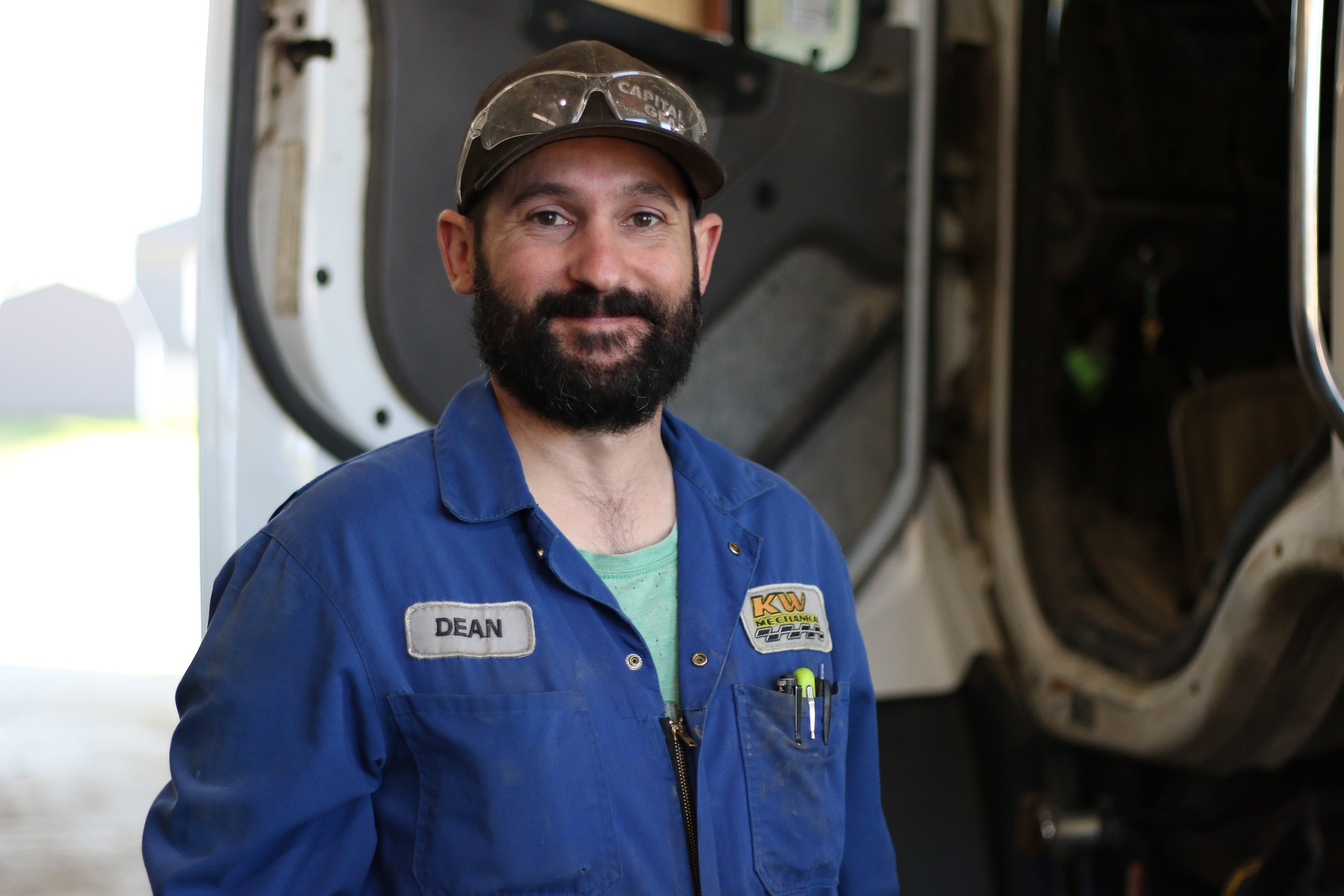 A man with a beard wearing a blue mechanic's jumpsuit and a cap, standing inside an auto repair shop with a vehicle in the background.