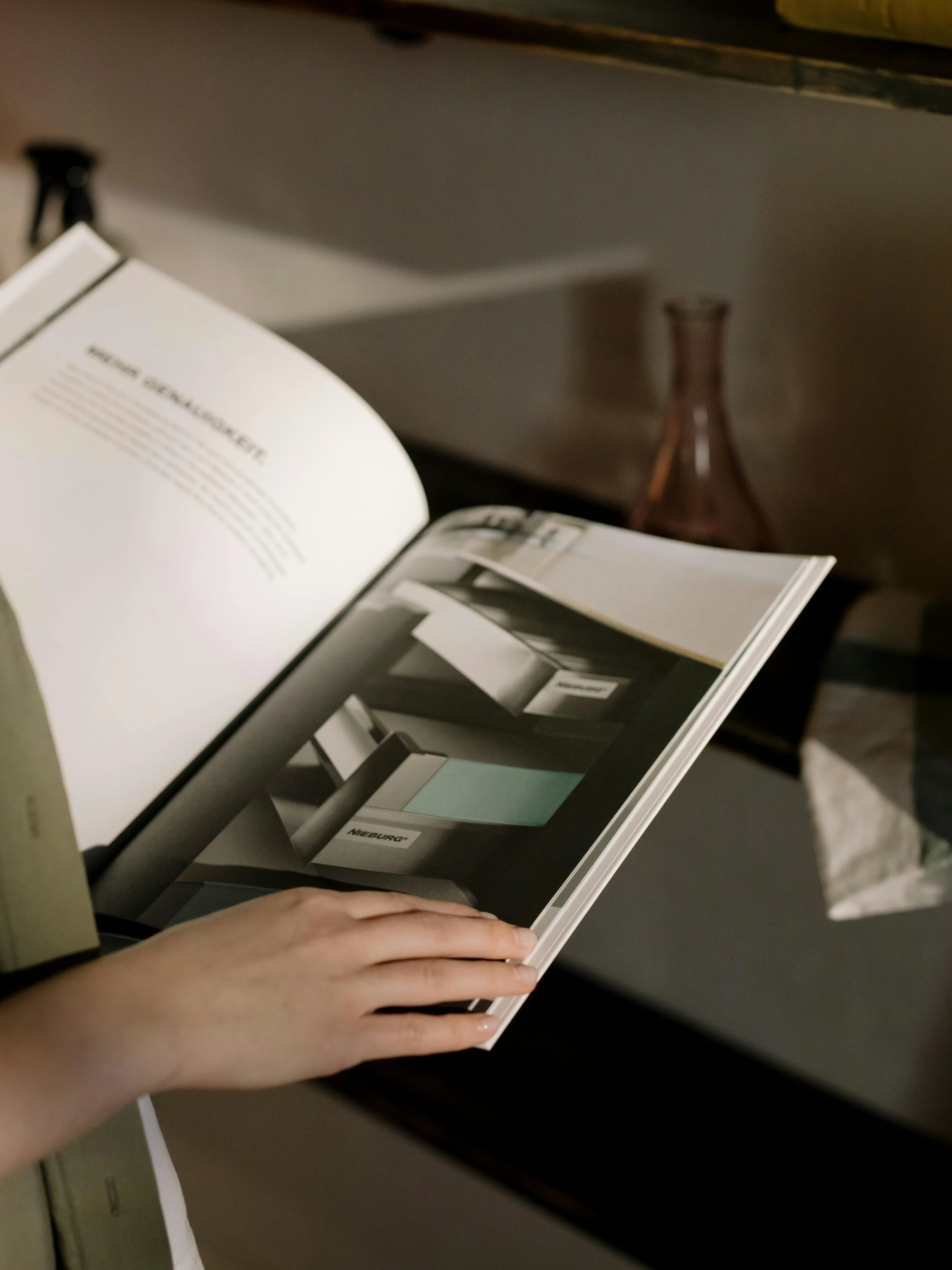 Person holding an open magazine with interior design images, sitting at a desk with a computer monitor and a vase in the background.