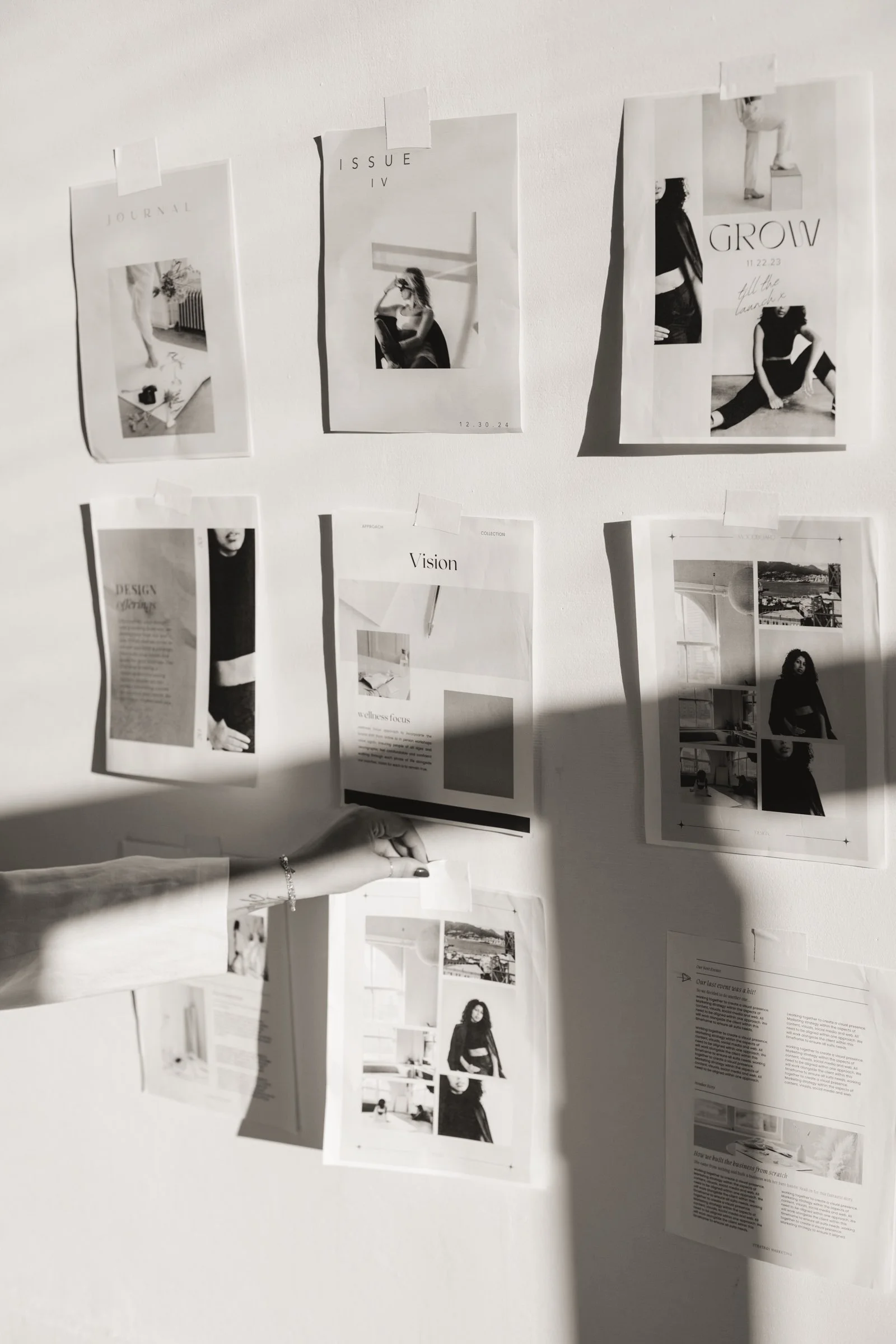 Black and white photo of a wall with various printed pages and photographs taped to it, with a person’s hand and arm reaching out to adjust one of the pages, shadows cast on the wall.