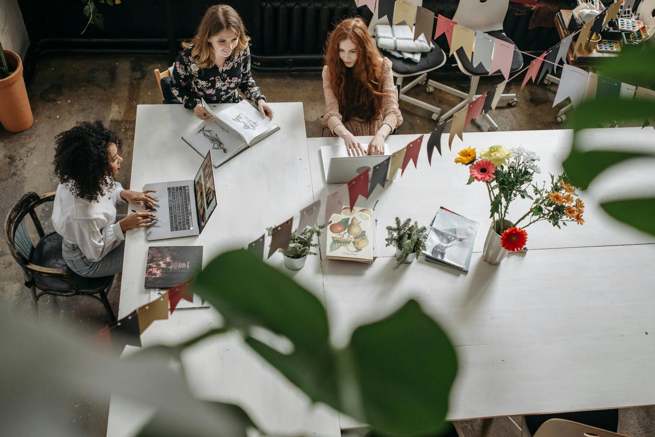 Three women working around a white table with plants, books, a laptop, and decorations in a creative workspace.