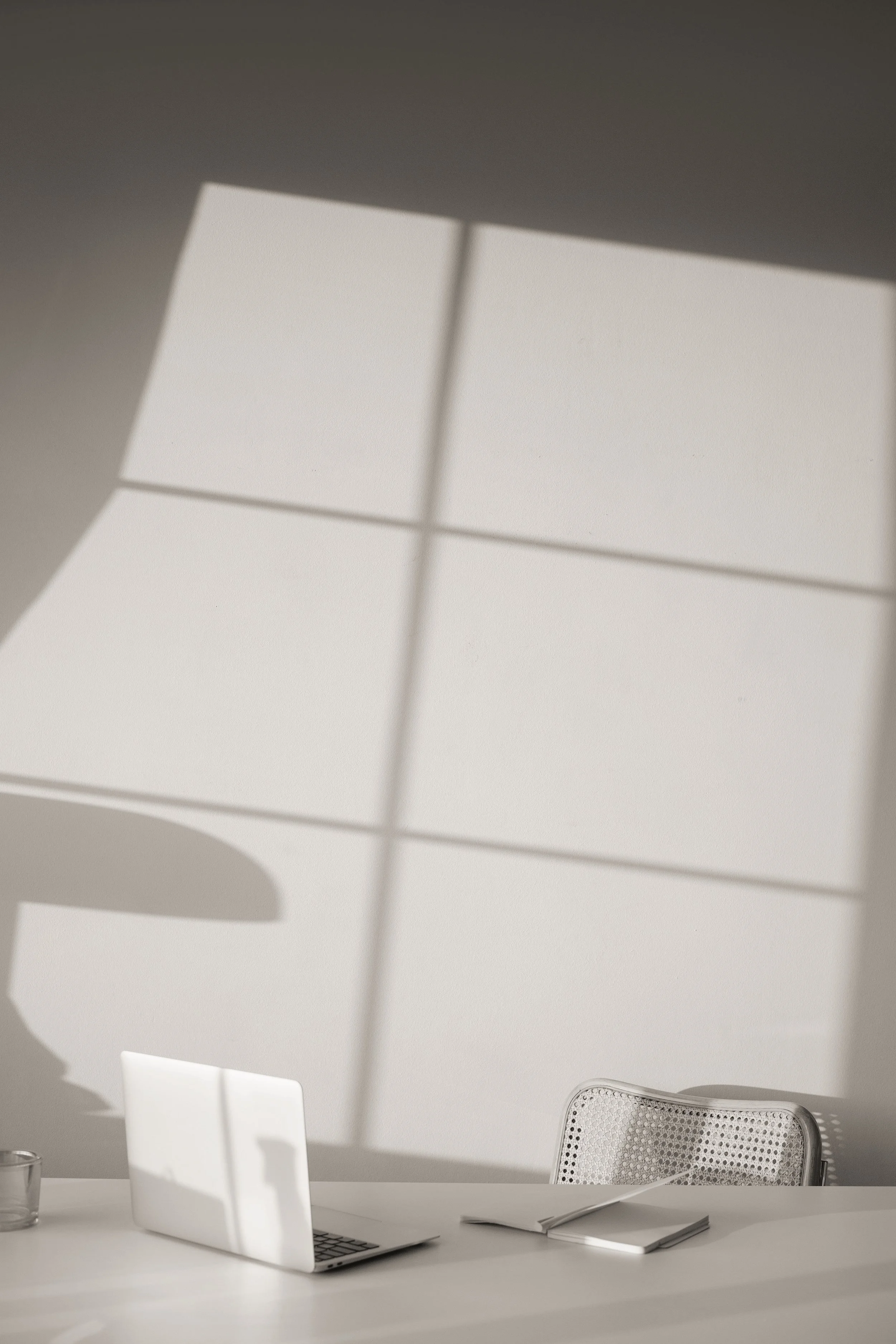 Sunlight casting a grid shadow on a white desk with a silver laptop, a glass, a closed notebook, and a chair with a woven backrest.