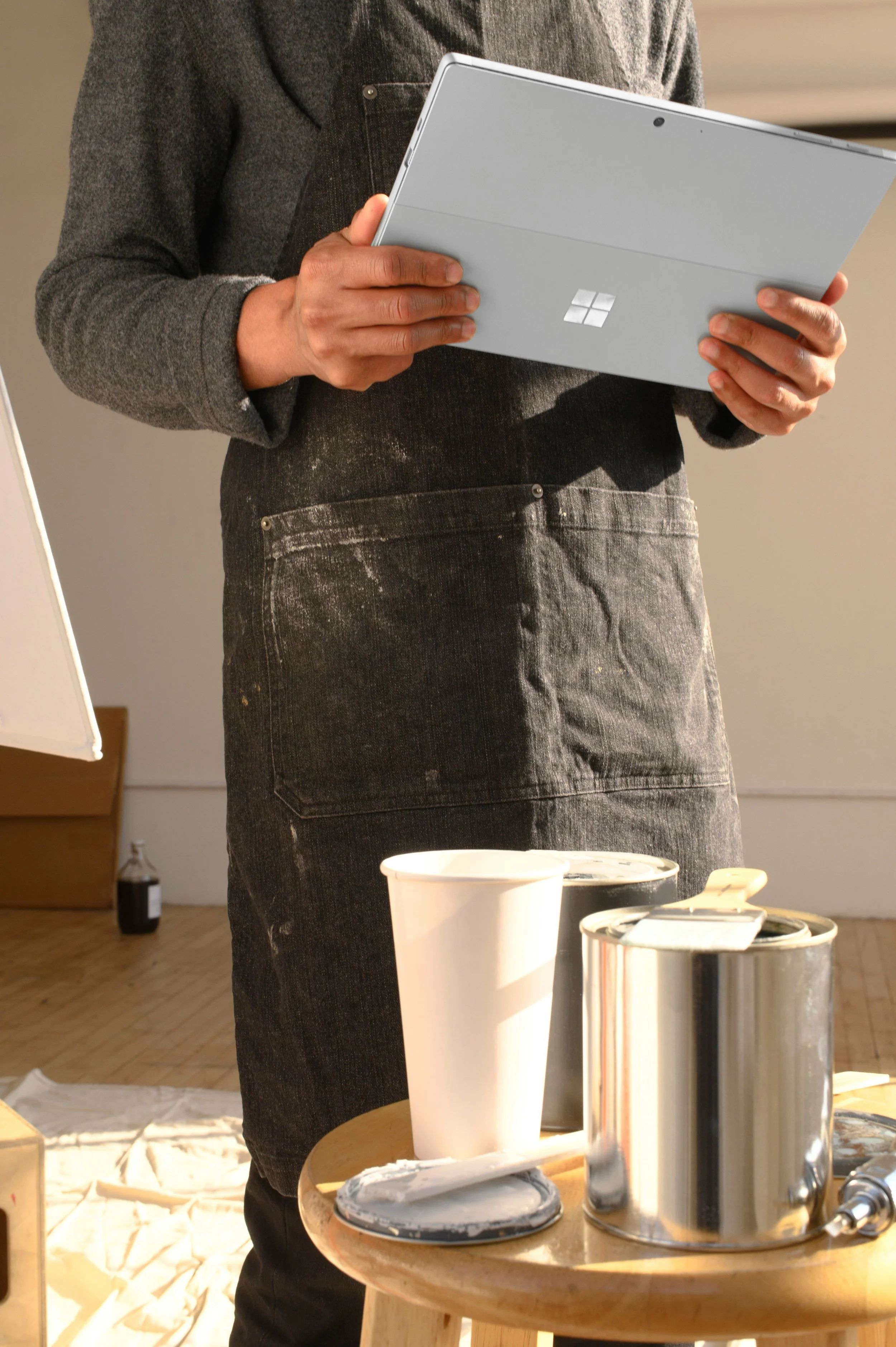 Person wearing a black apron holding a silver Microsoft Surface tablet, with paint supplies on a nearby small wooden table.
