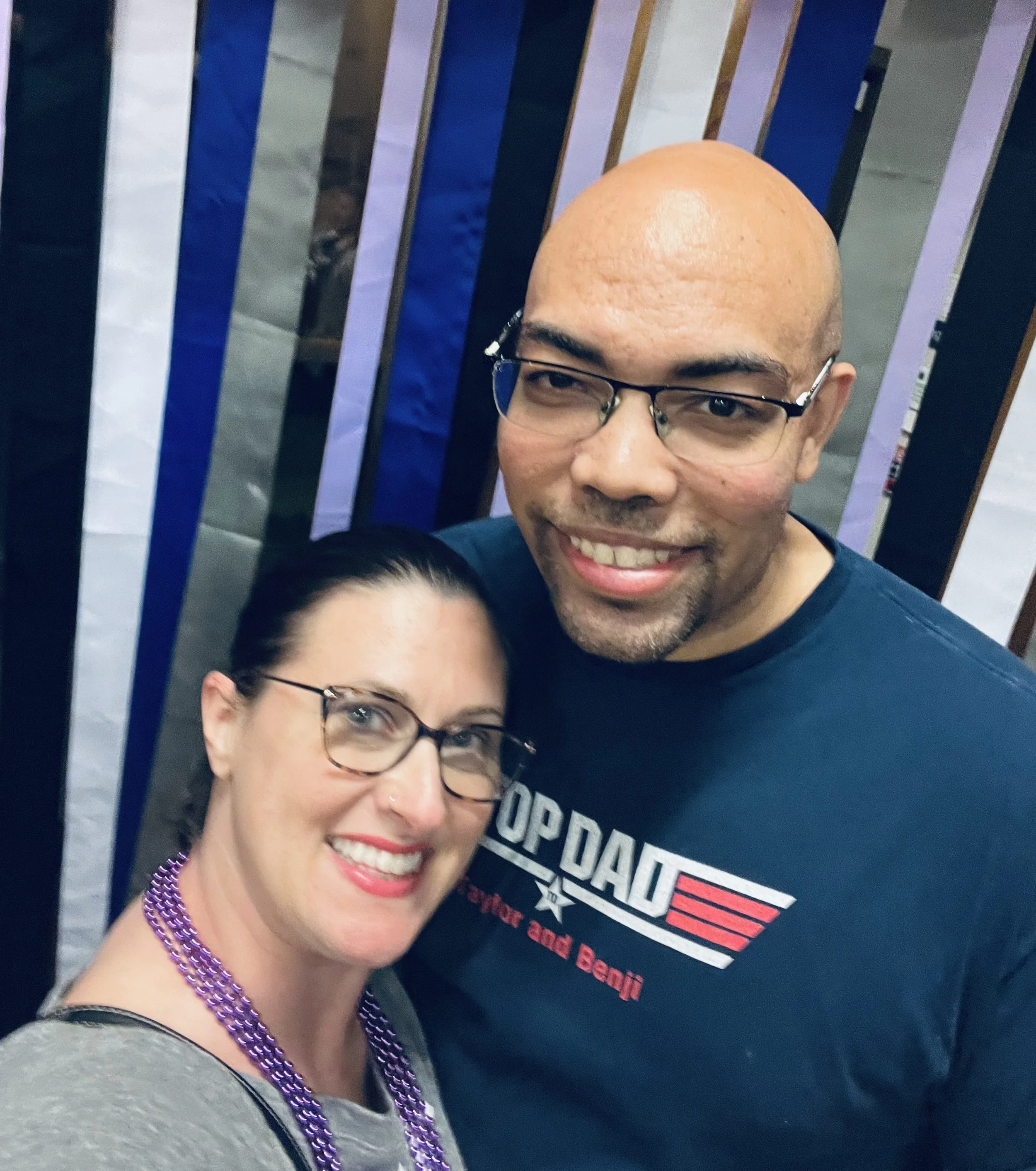 The owners, a woman and a man smiling for a selfie, both wearing glasses, standing in front of a colorful striped background.