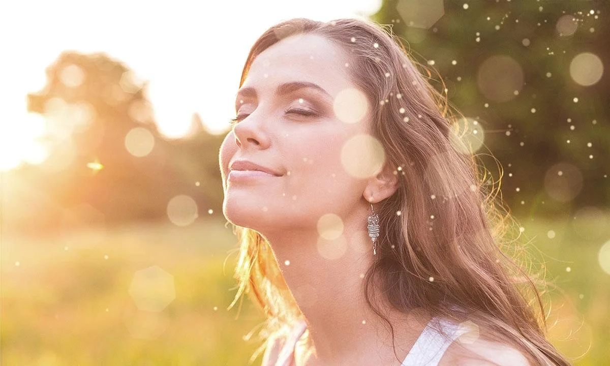 A woman with long brown hair, closed eyes, and a gentle smile standing outdoors in warm sunlight, with a blurred natural background and sparkling bokeh effects.