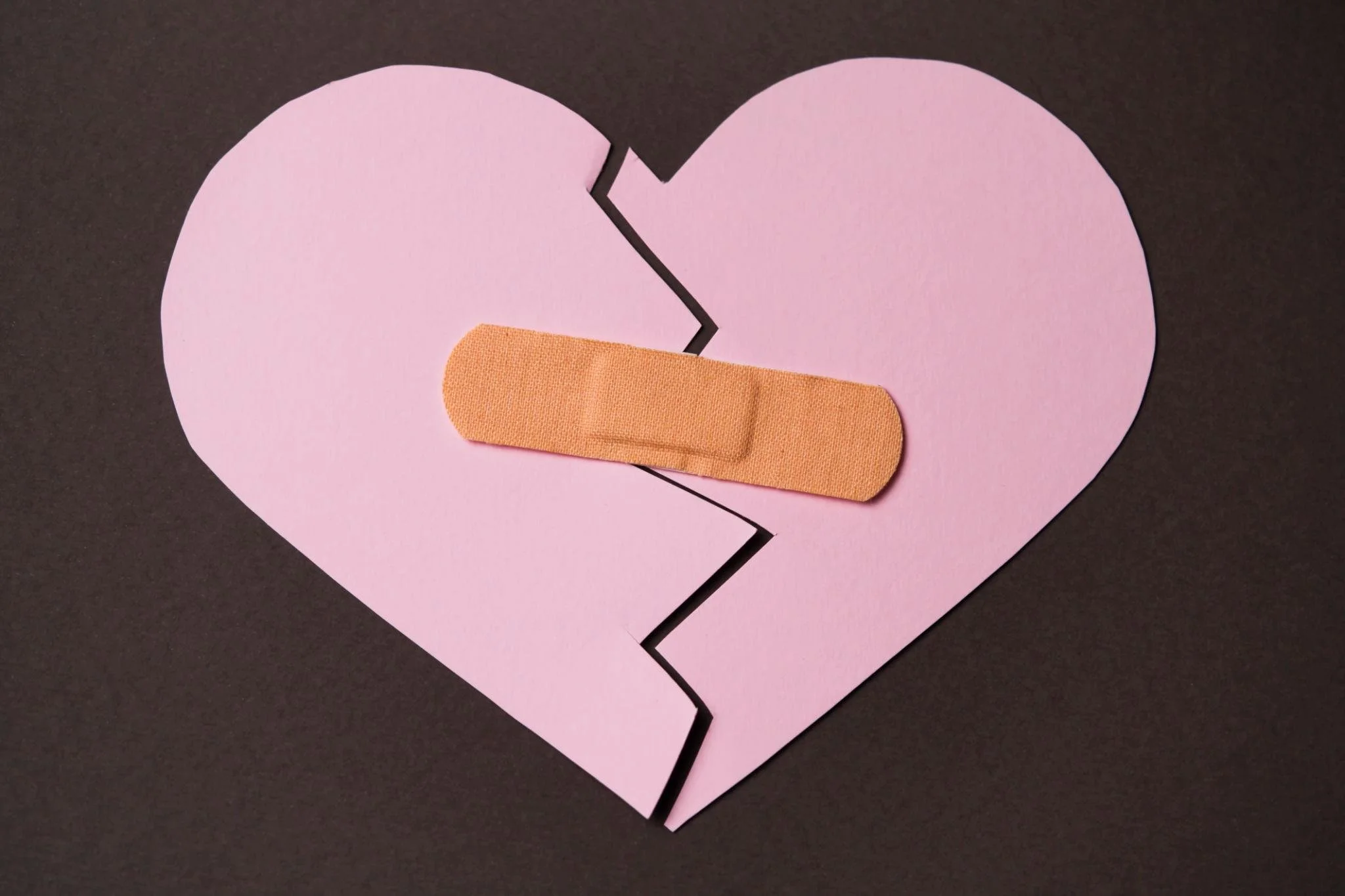 A pink paper heart with a black crack running down the middle, and a beige adhesive bandage placed over the crack, on a dark background.