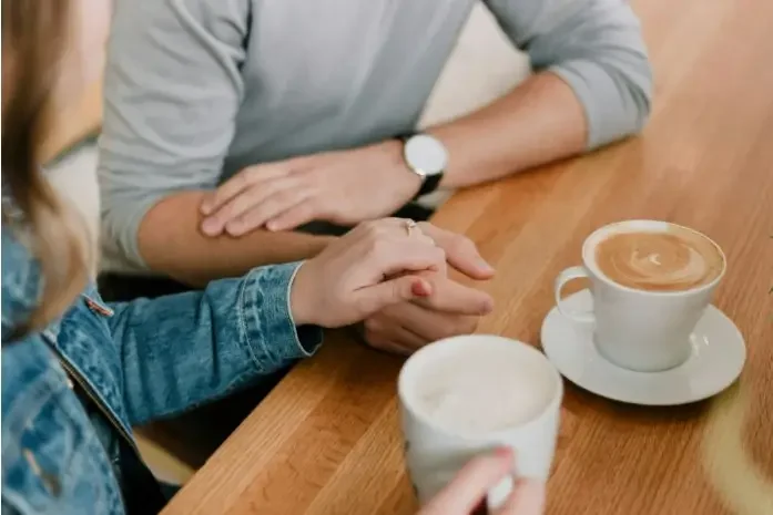 A couple holding hands at a coffee shop, with one partner appearing distant and emotionally unavailable.