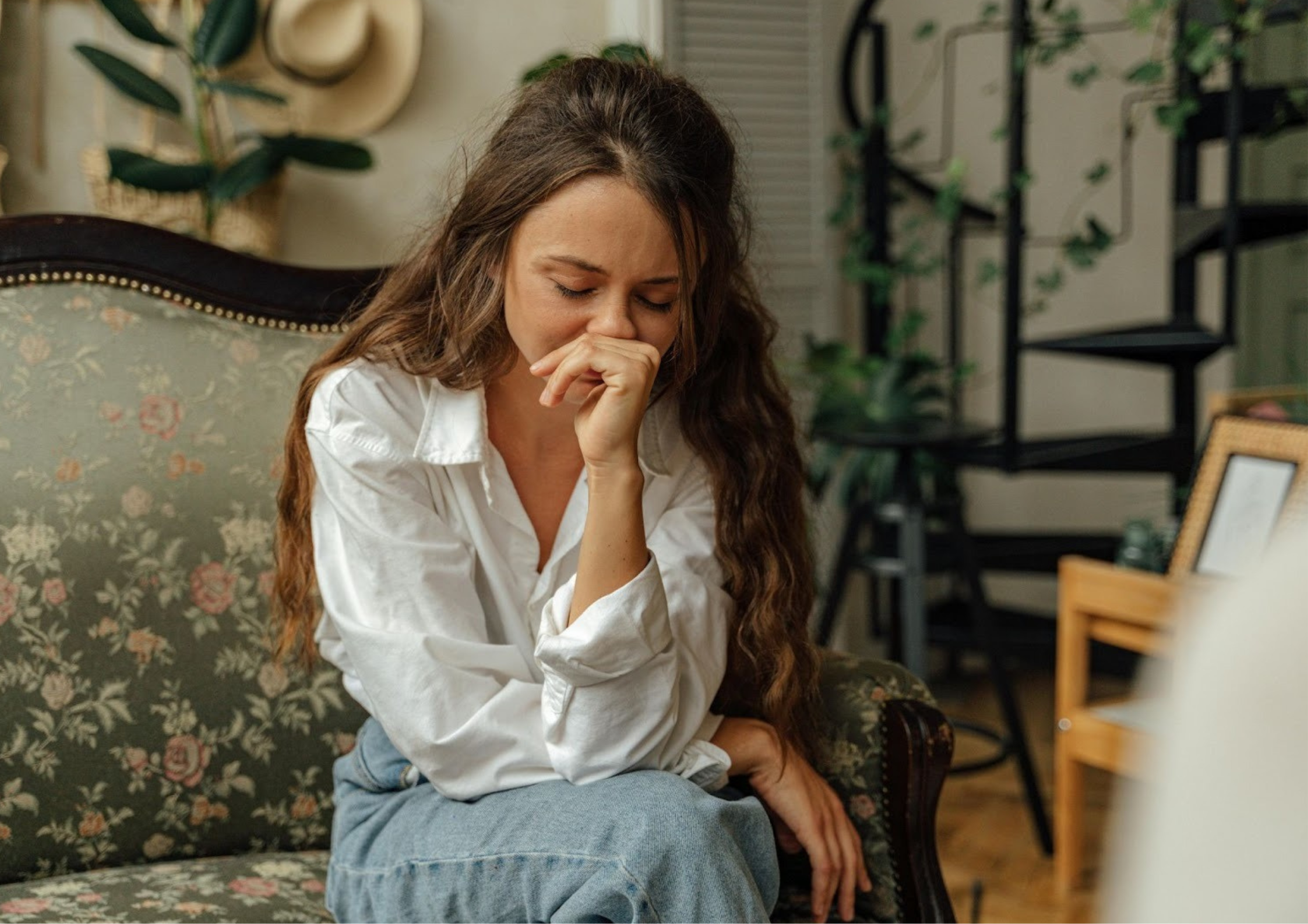 A woman sitting on a floral-patterned couch with her head bowed and hand to her face, appearing distressed or deep in thought in a therapist’s office.