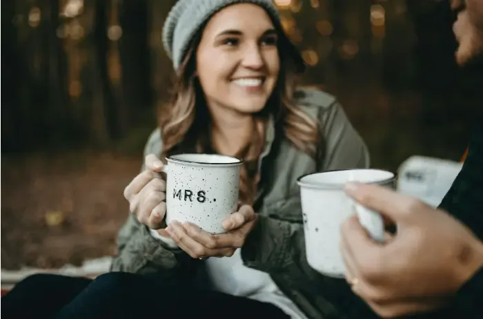 A couple in the woods, holding mugs and enjoying a cozy moment together amidst the trees.