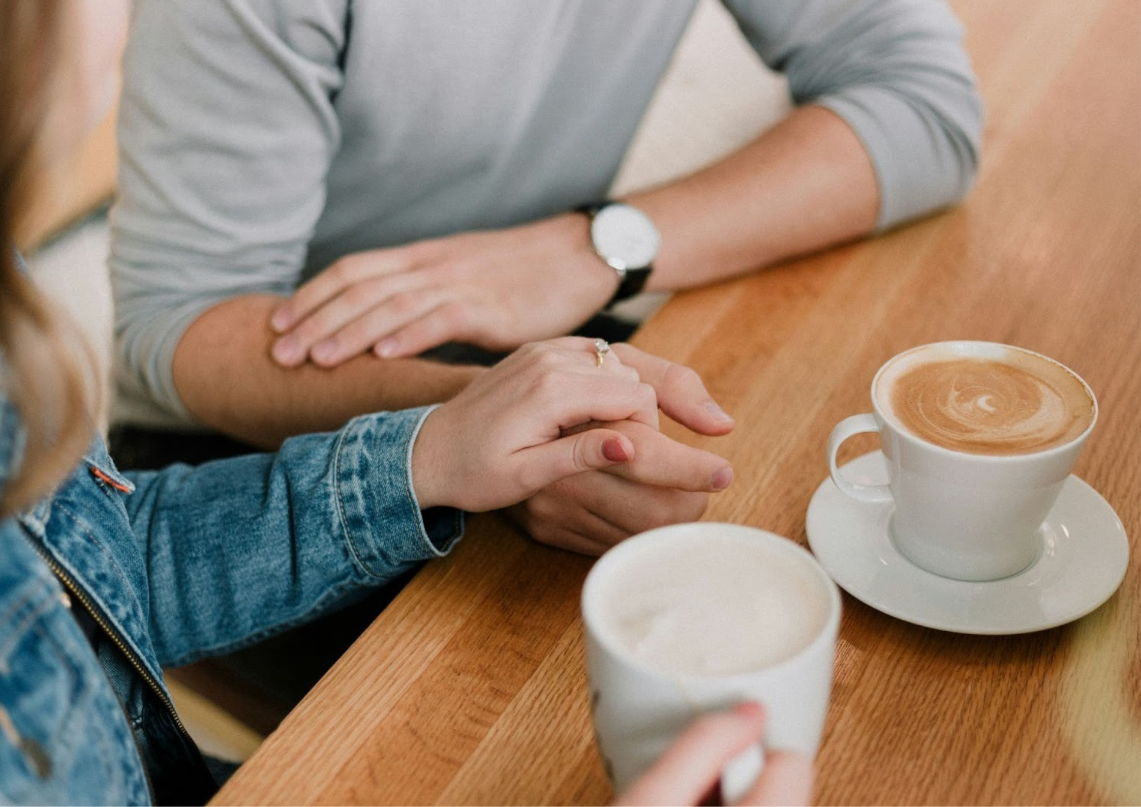 A close-up of two people sitting at a table with coffee cups, gently holding hands in a supportive or intimate gesture.