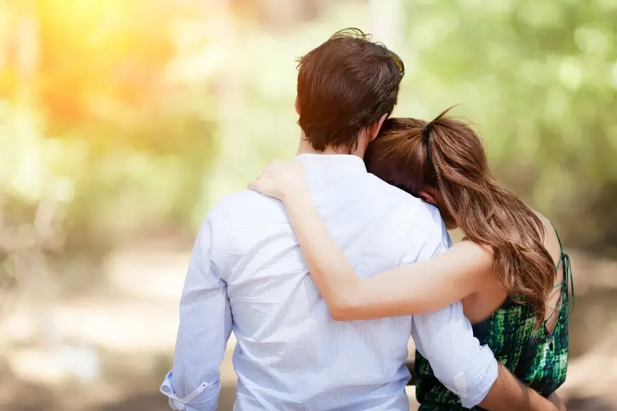 Couple embracing outdoors, woman resting her head on man's shoulder, with warm sunlight and greenery in the background.