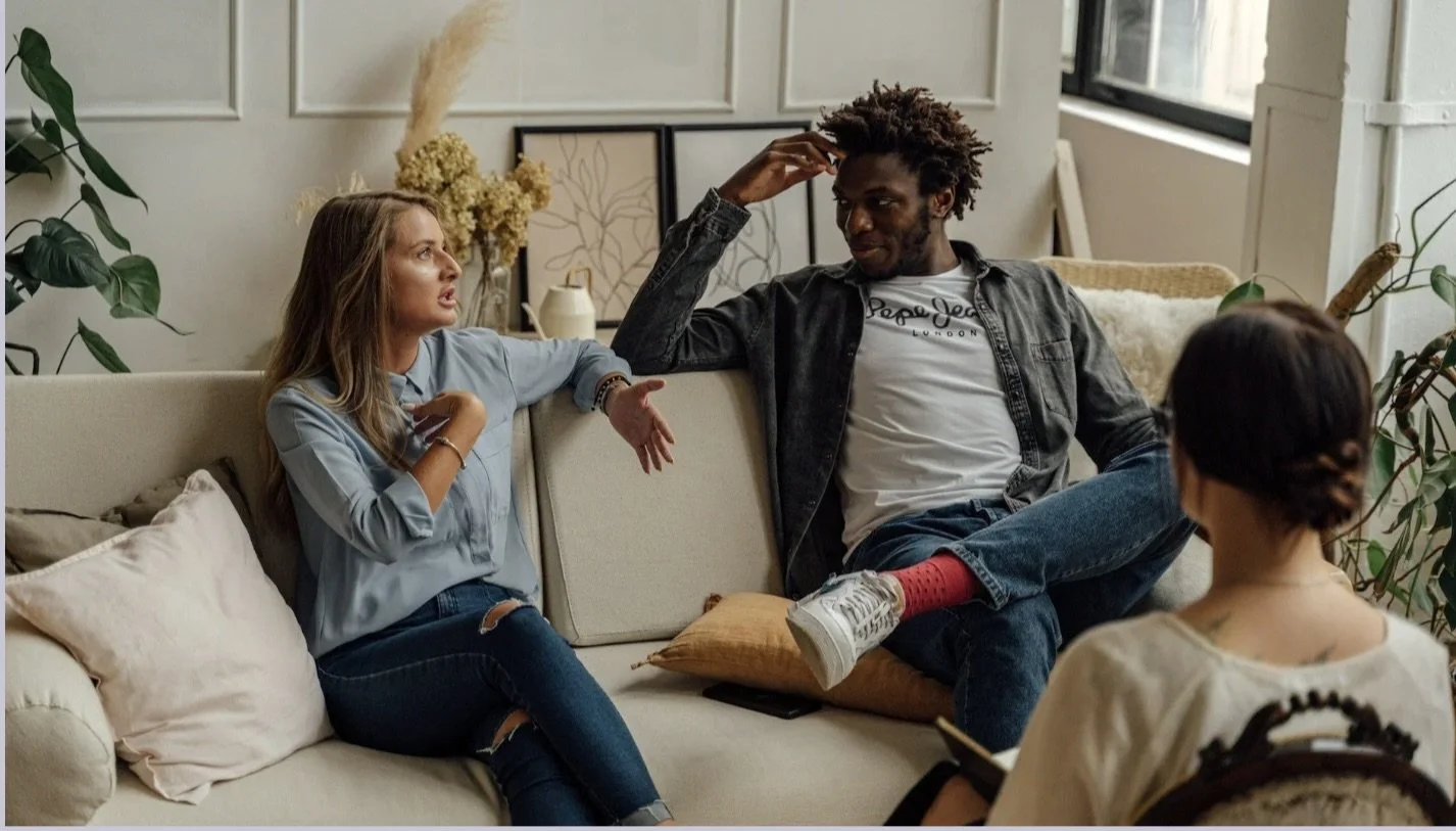 Three people having a conversation on a beige couch in a cozy, well-decorated living room.