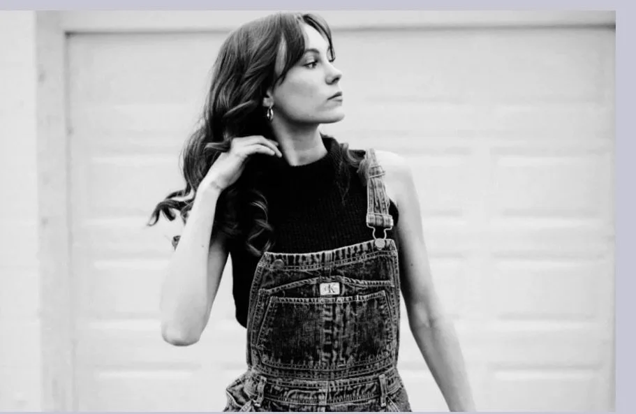A black and white photo of a woman with long, wavy hair, wearing a sleeveless top and denim overalls.