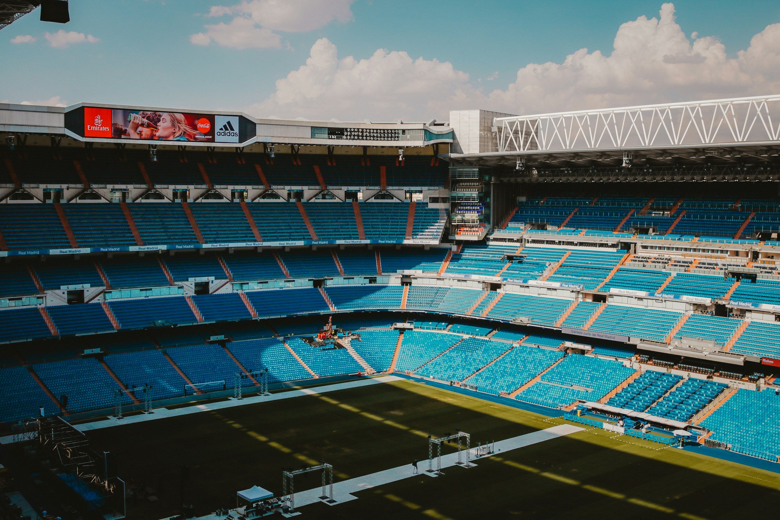 Empty soccer stadium with blue seats, a green field, and a bright sky with clouds.