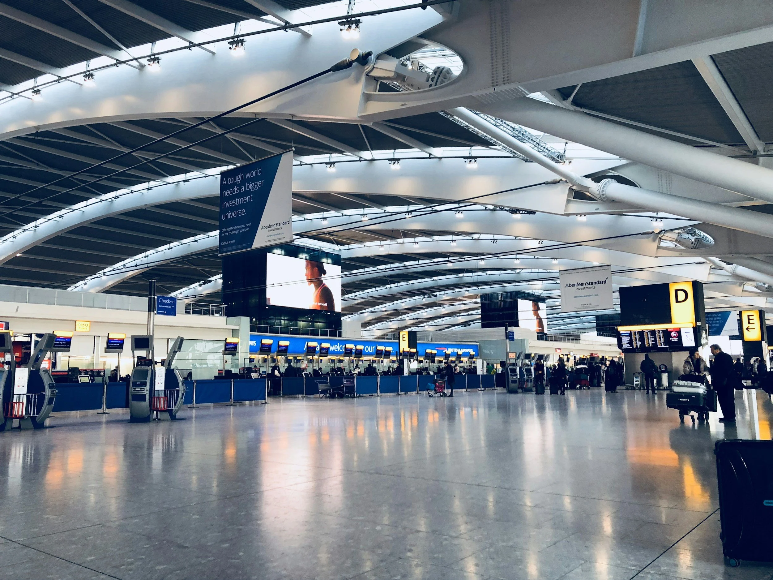 Empty airport check-in counters and seating area with passengers and luggage, large digital screens, and overhead signage.