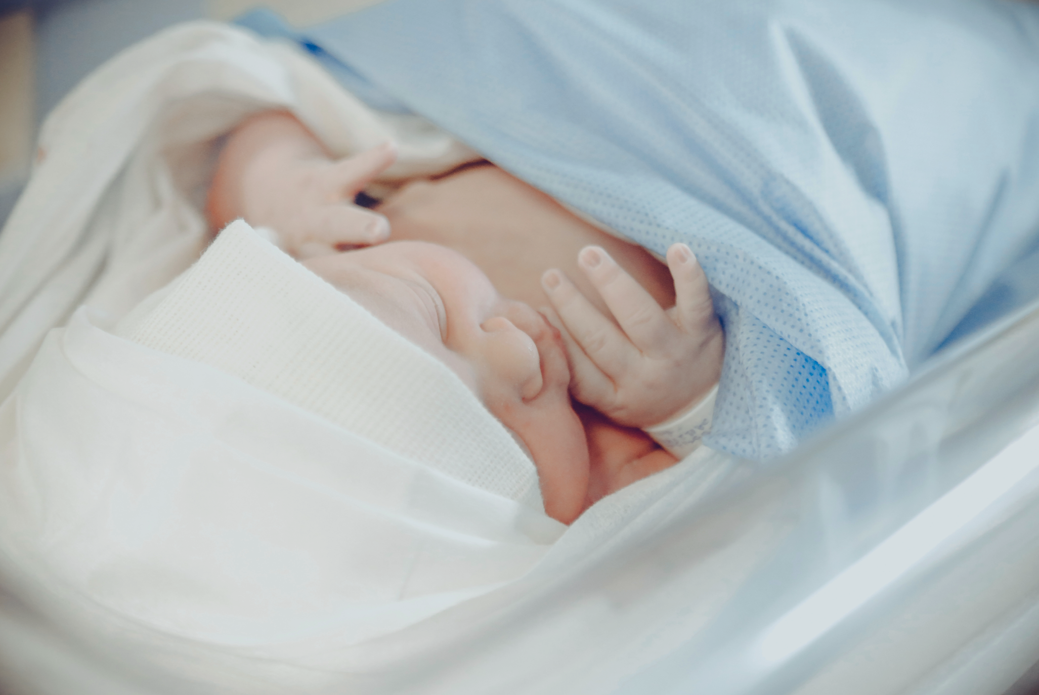 Newborn baby wearing a white hat and sleeping, lying in an incubator, with hand touching face, covered by a blue hospital blanket.