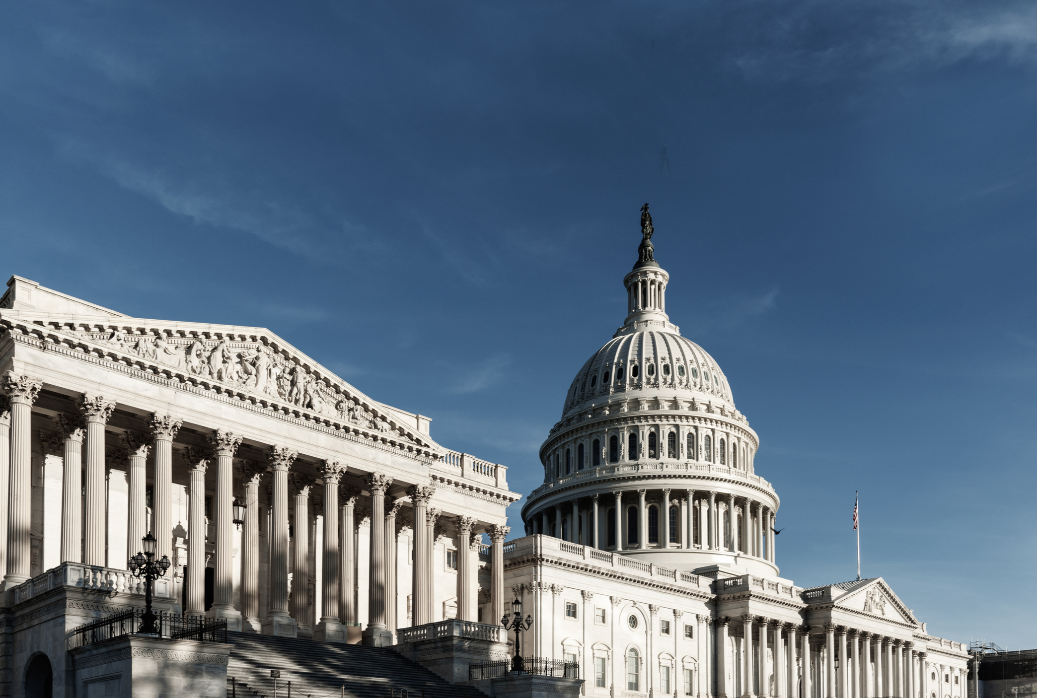 The United States Capitol building with a prominent dome, columns, and a flag flying on the right, under a clear blue sky.