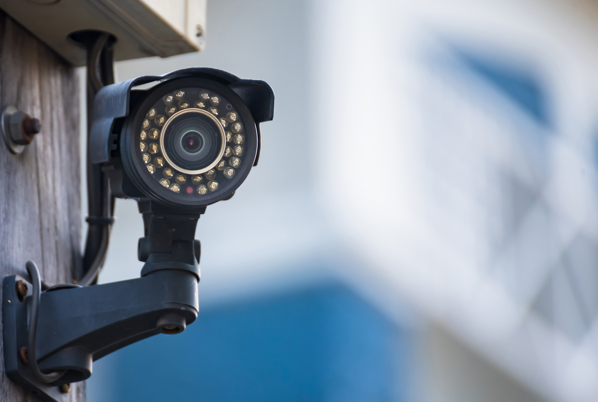 Close-up of a security camera mounted on a wooden pole with a blurred background.