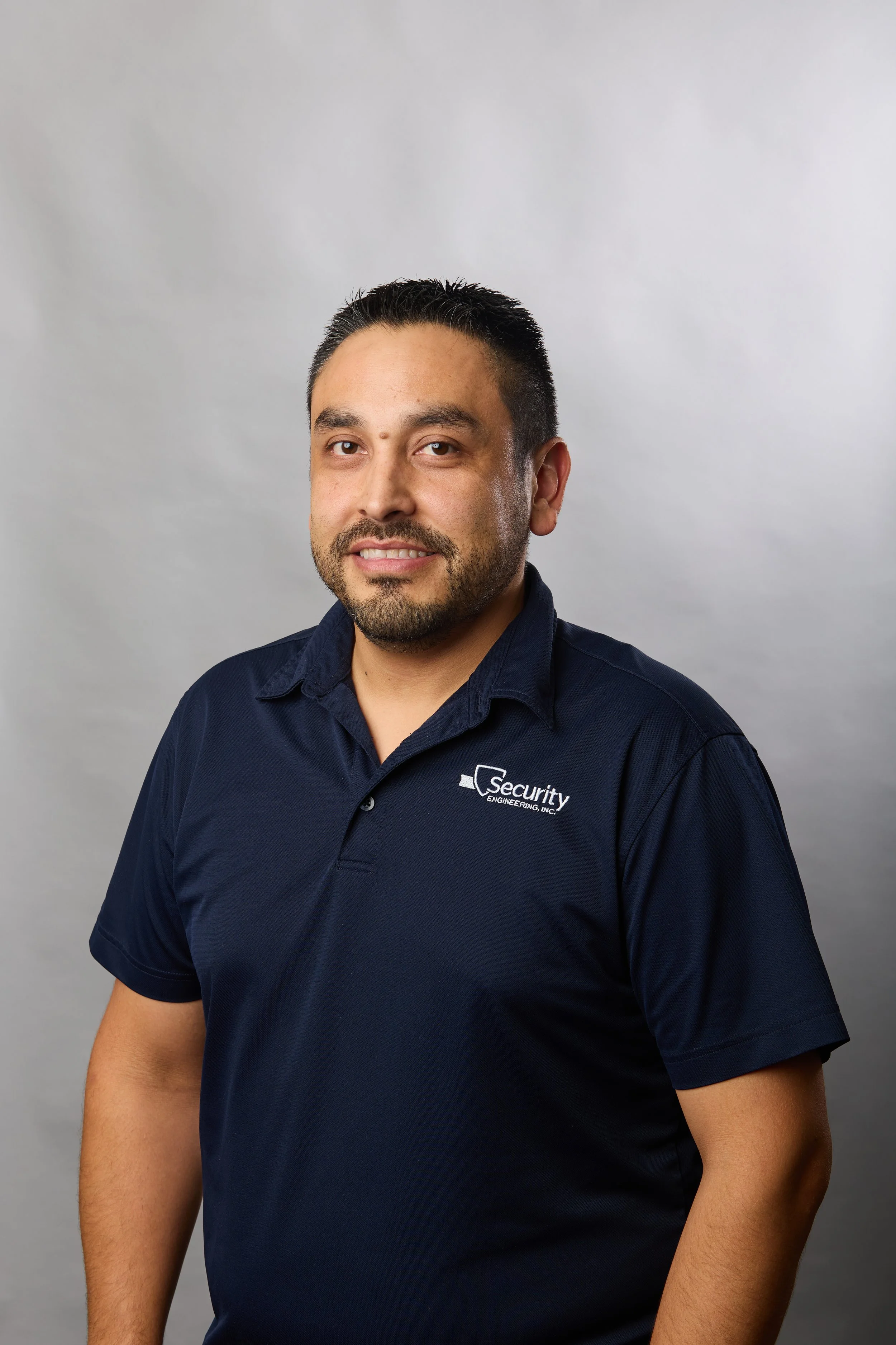 Portrait of a man in a navy blue polo shirt with the logo 'Security Engineering Inc.' on the chest, standing against a light gray background.