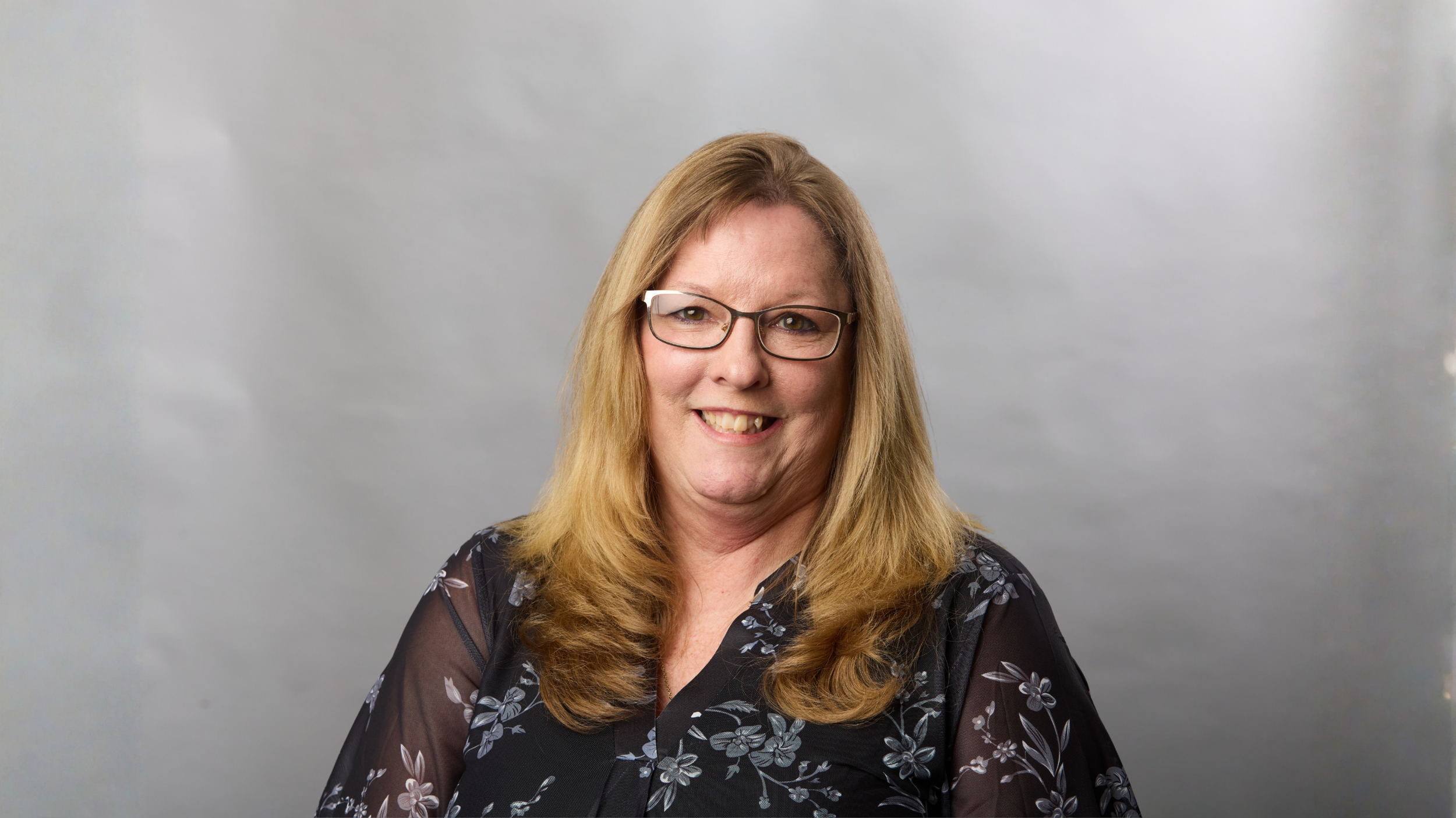 A middle-aged woman with light skin, long blonde hair, glasses, and a black floral blouse, smiling against a plain grey background.