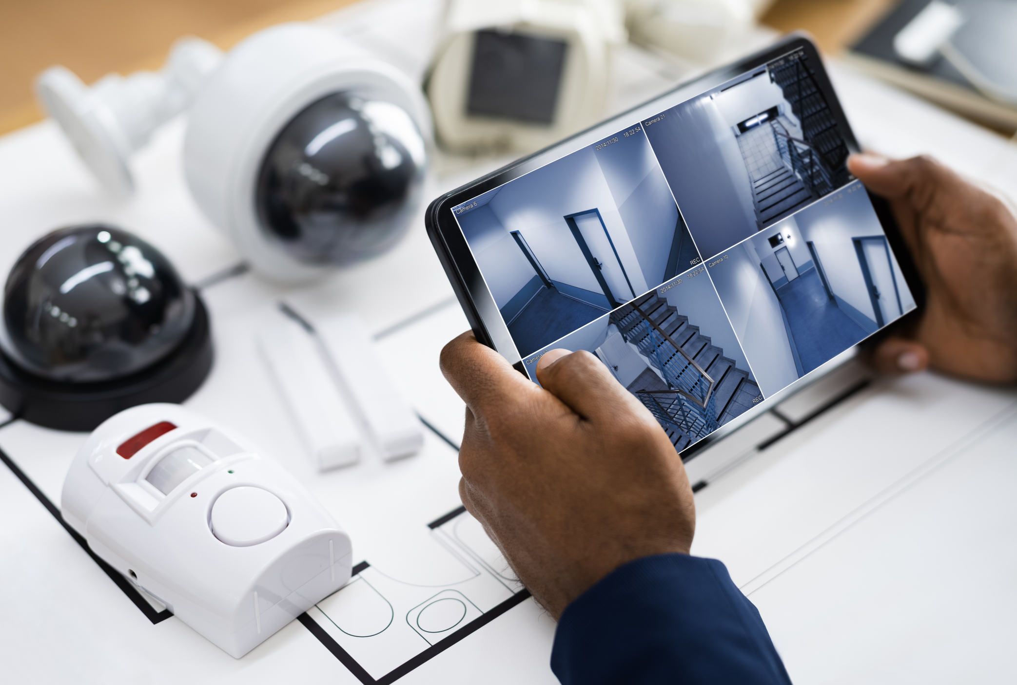 Person holding a tablet displaying security camera footage of stairs, hallway, and doorways, with security cameras and smoke detectors on a planning table.