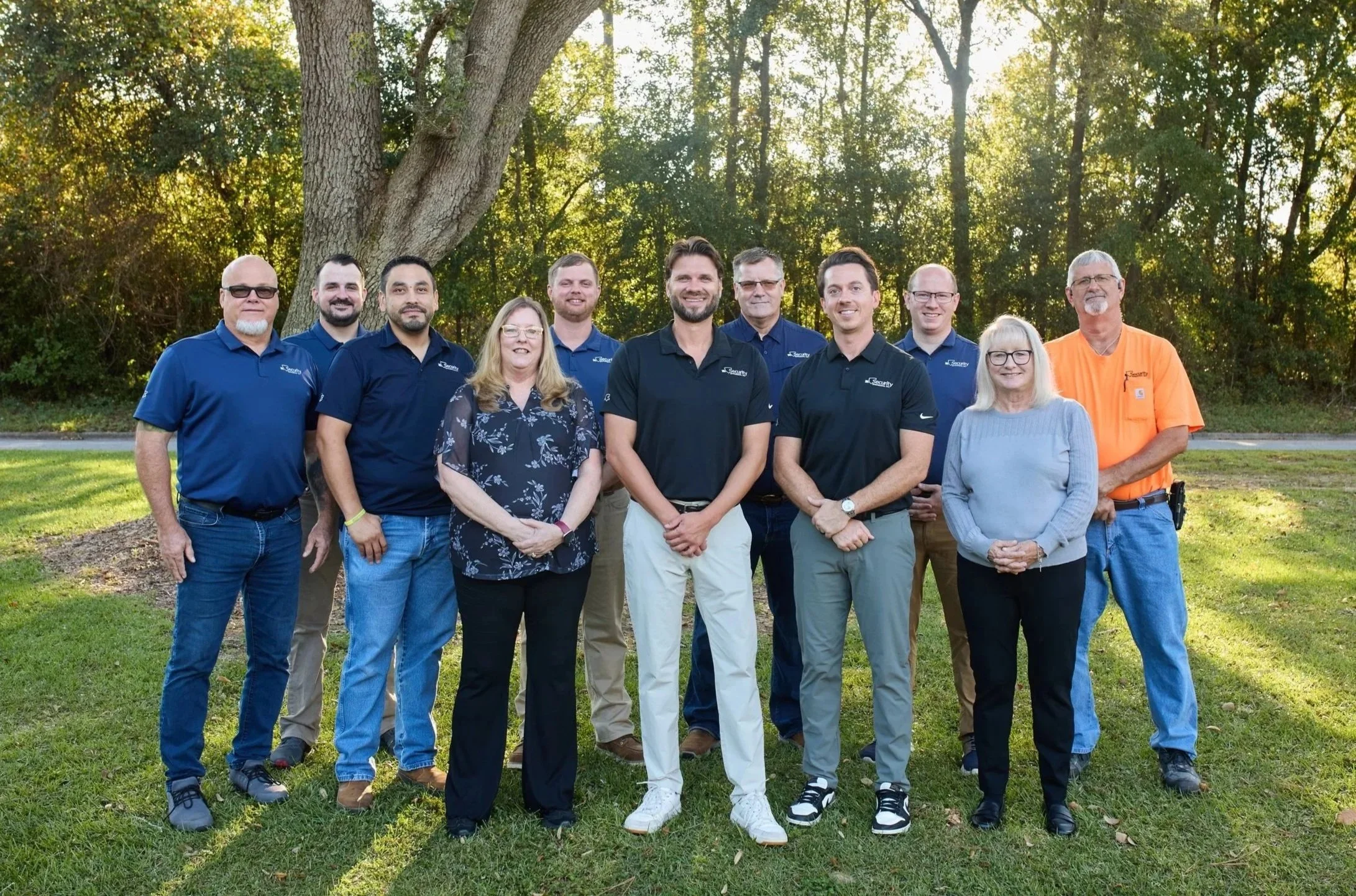 Group photo of eleven people standing outdoors on grass in front of trees, dressed in casual and business casual attire, smiling at the camera.