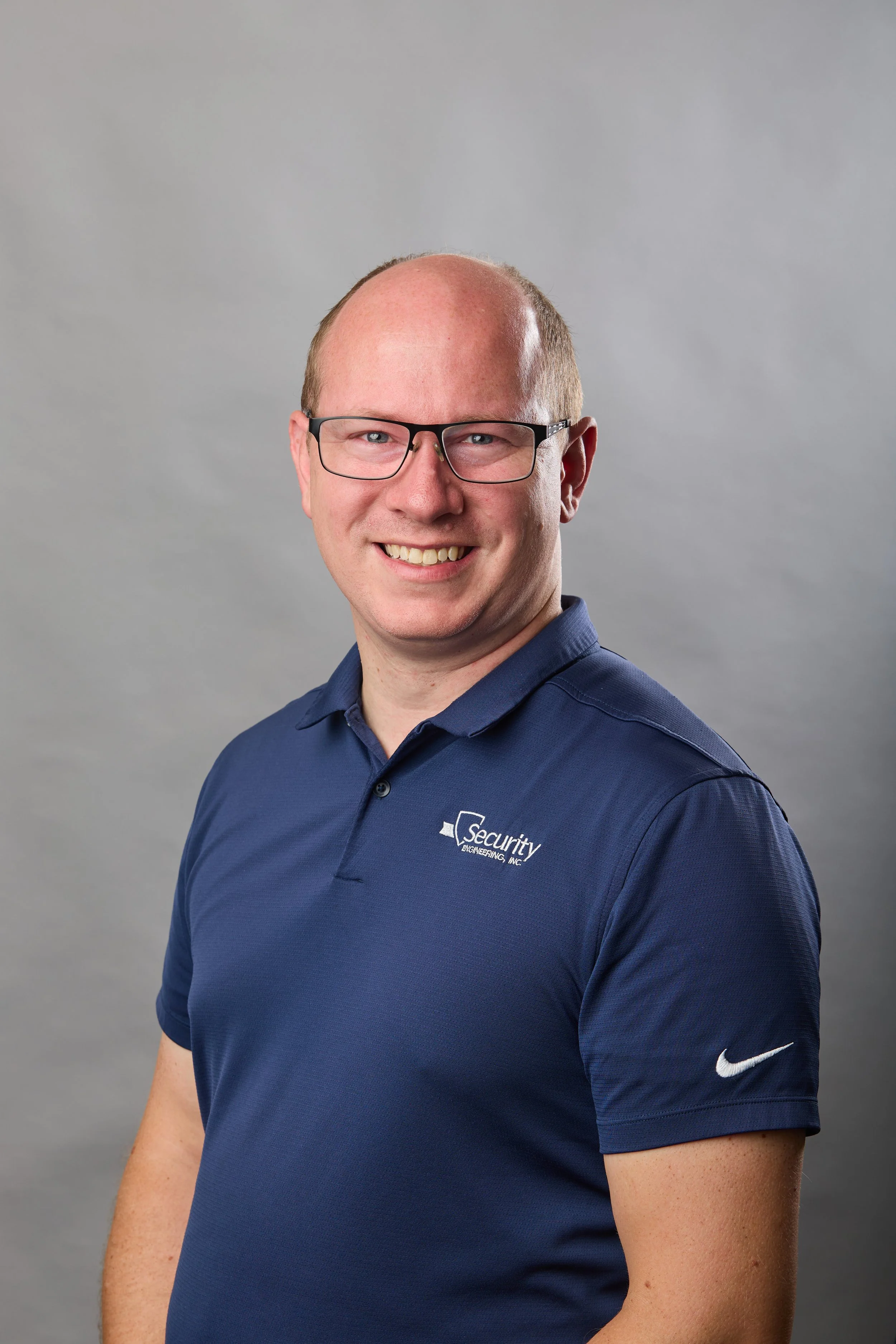Portrait of a smiling man with glasses, wearing a navy blue polo shirt with a company logo that reads "Security Engineering, Inc." and a white Nike logo on the sleeve, standing against a light gray background.