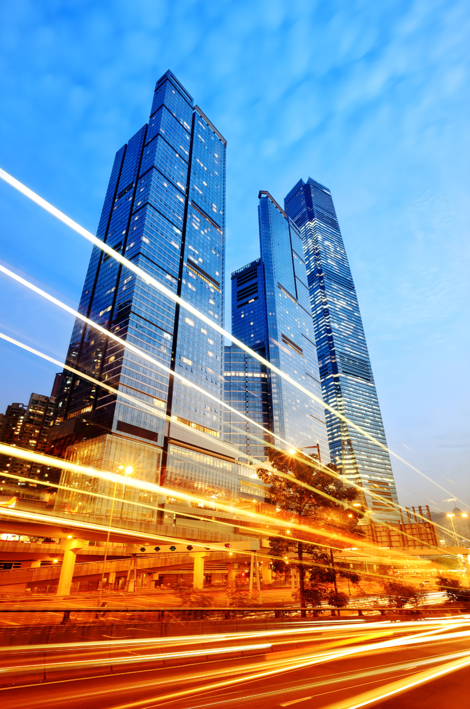 Tall modern glass skyscrapers in a city with light trails from moving vehicles in the foreground during dusk.