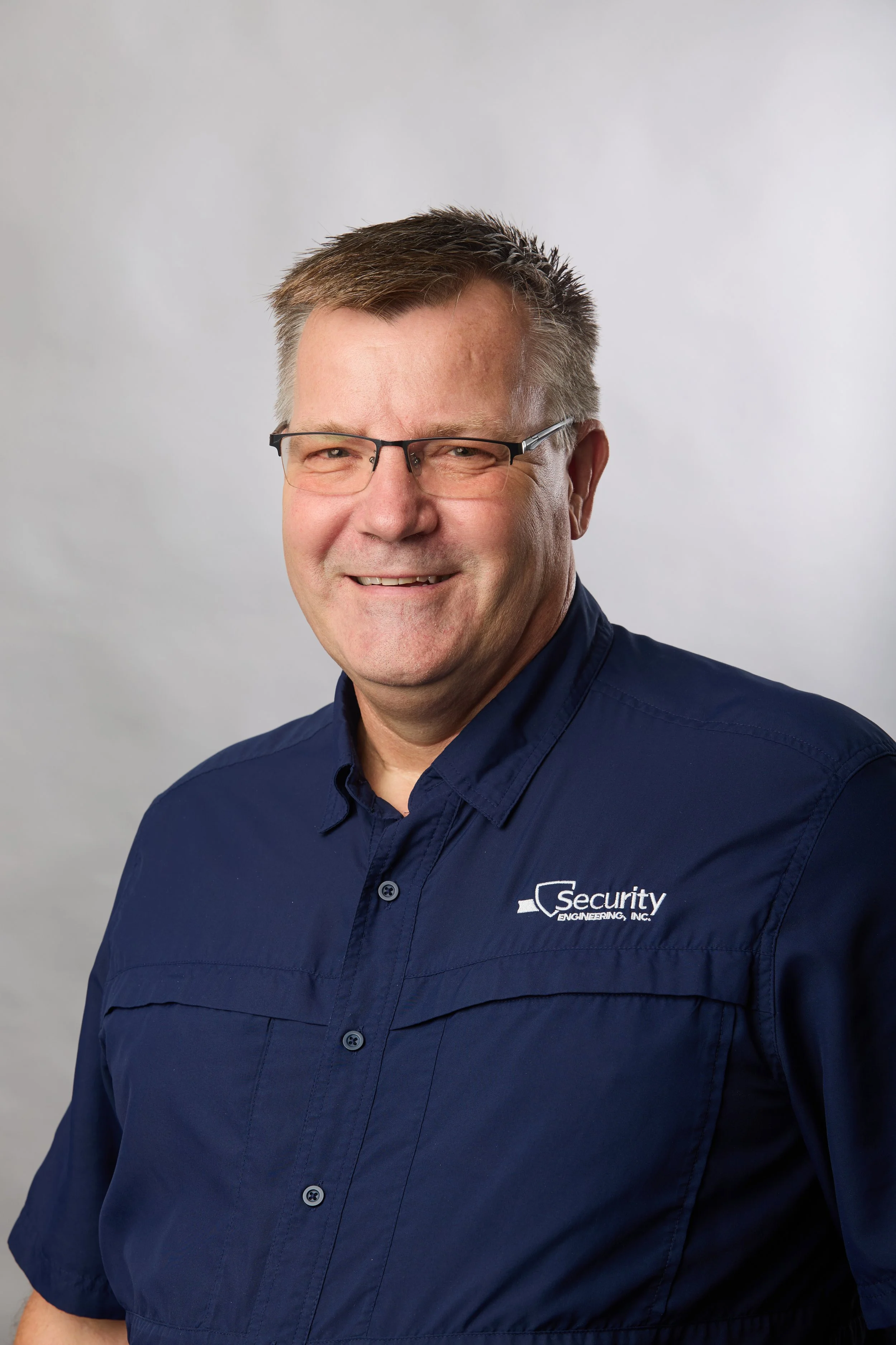 A smiling man with short brown hair and glasses wearing a navy blue shirt with a security logo.