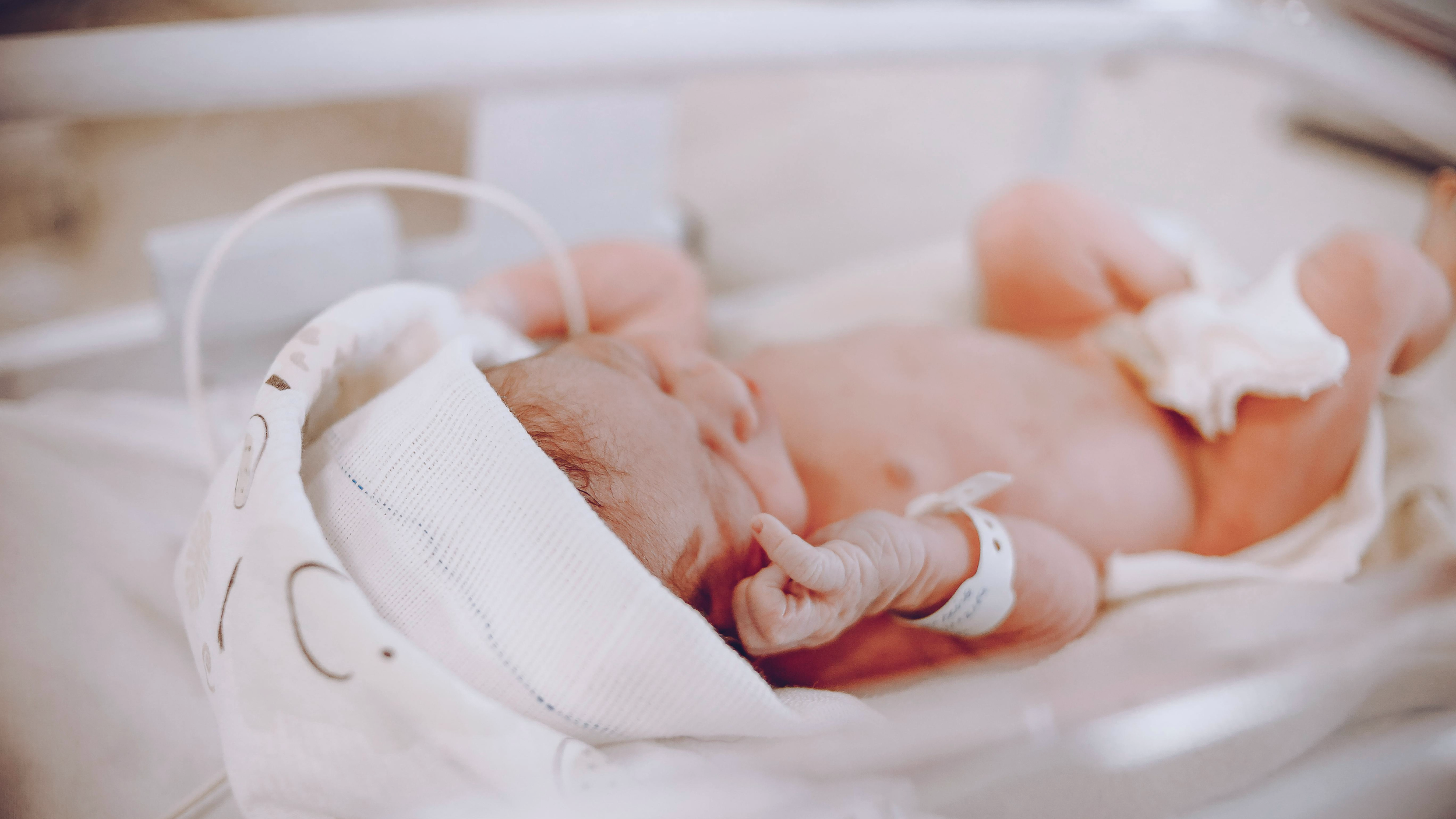 Close-up of a newborn baby lying in a hospital bassinet, wearing a white cap and a diaper, with a hospital identification bracelet on their wrist.
