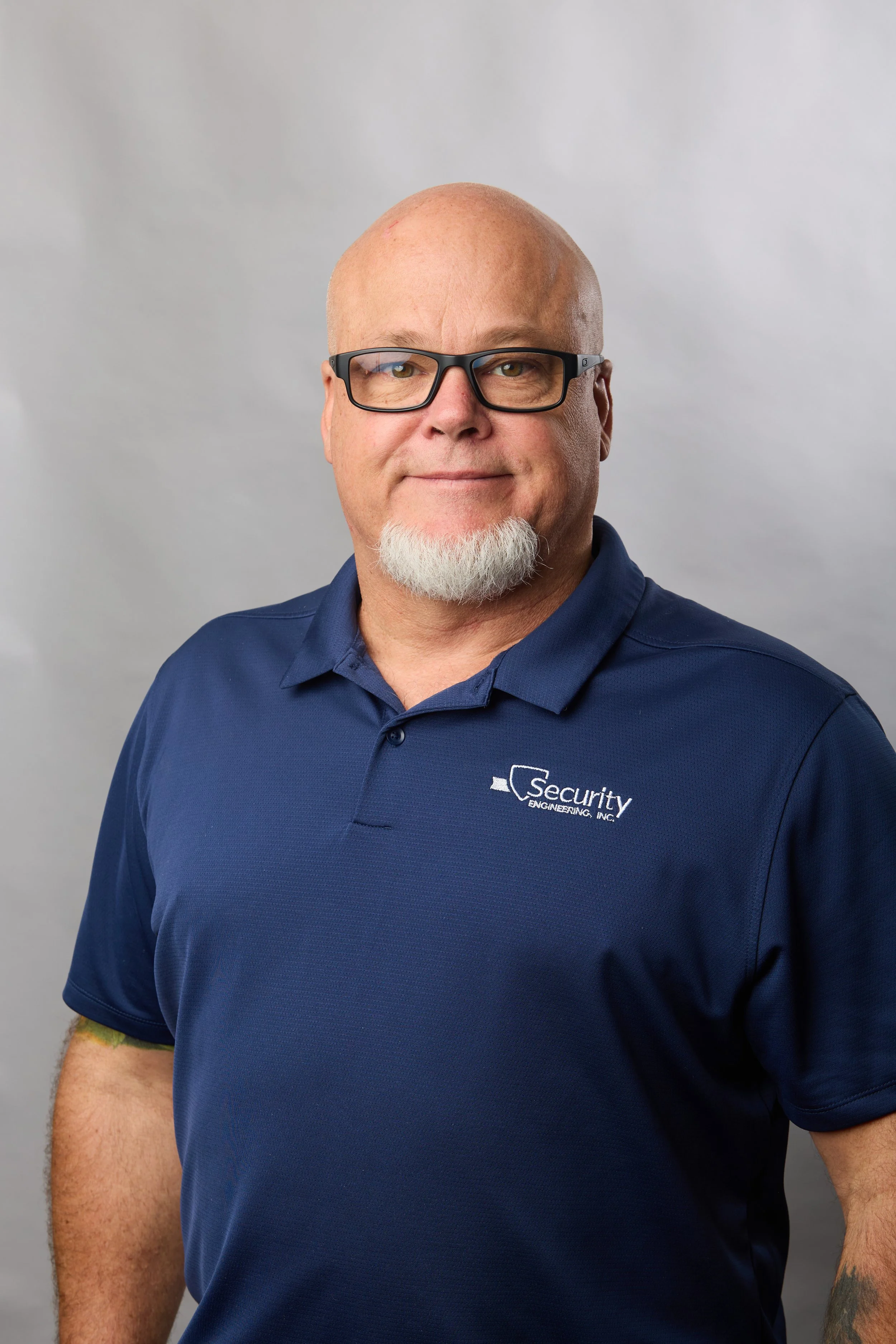 A man with glasses and a white goatee, wearing a blue polo shirt with a logo that says Security Engineering, Inc., standing against a plain background.