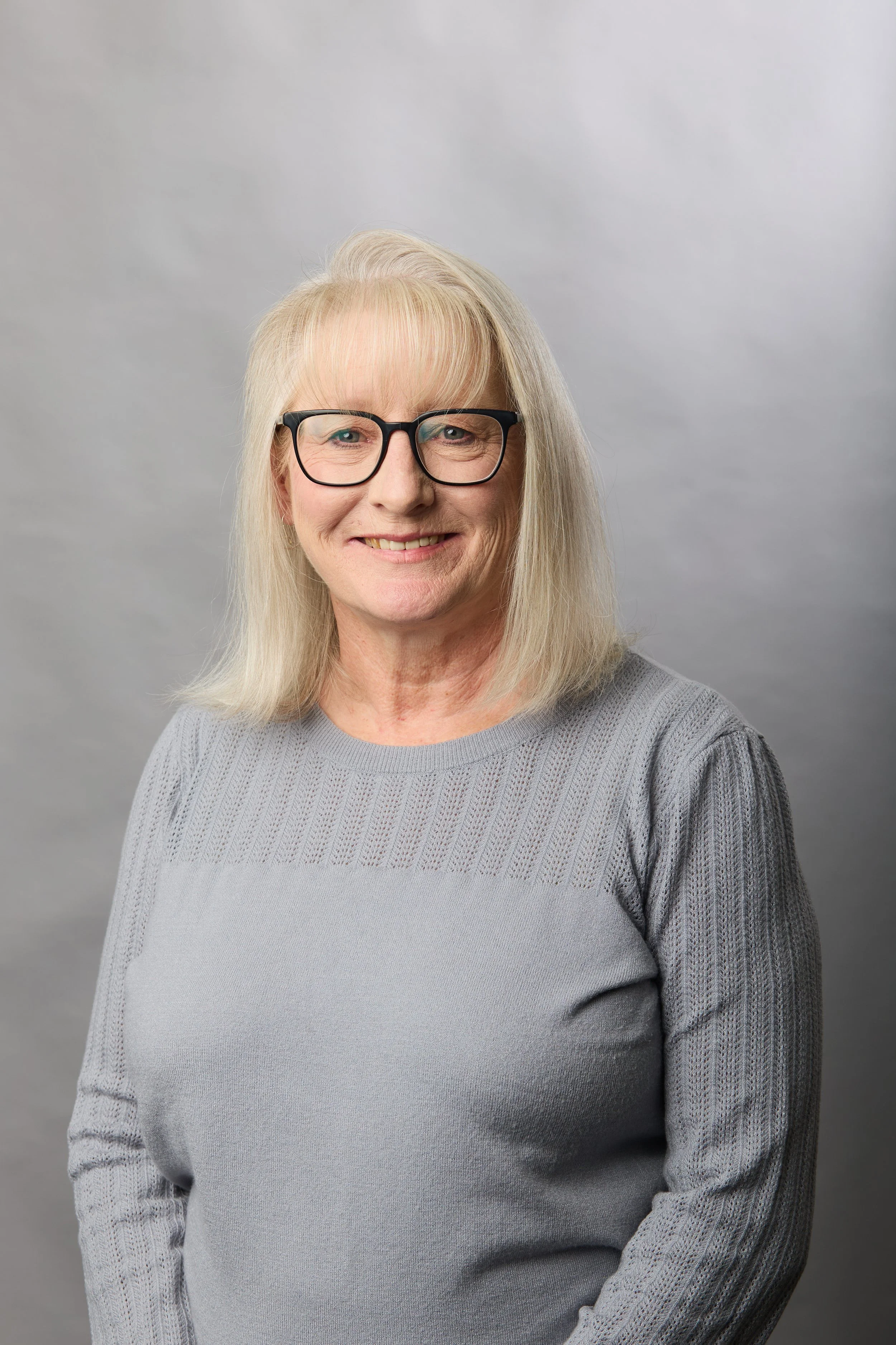 A portrait of a smiling middle-aged woman with blonde hair, wearing glasses and a light gray sweater, against a light gray background.
