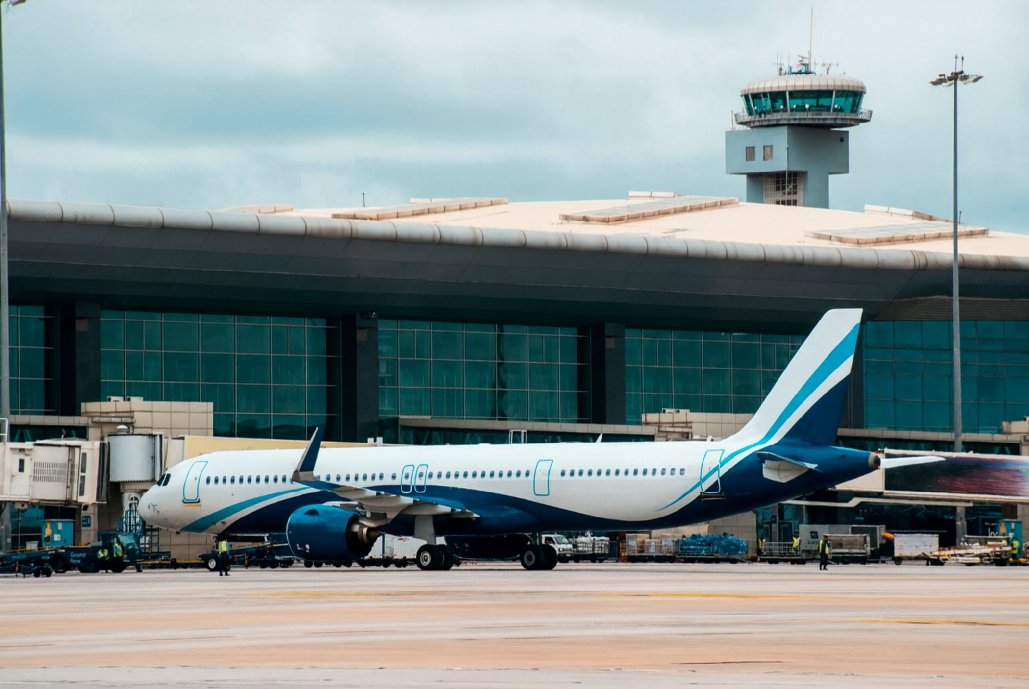 Commercial airplane parked at airport gate with jet bridge, airport tower in background, and ground crew working nearby.