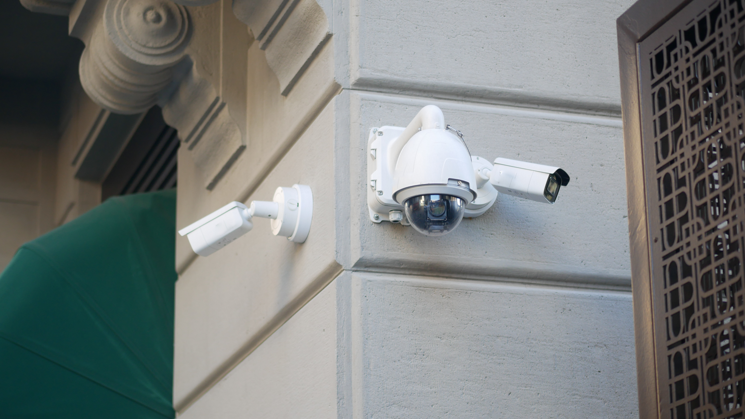Two security cameras mounted on the corner of a beige building, one with a dome and one with a rectangular shape, near a decorative brown metal structure.