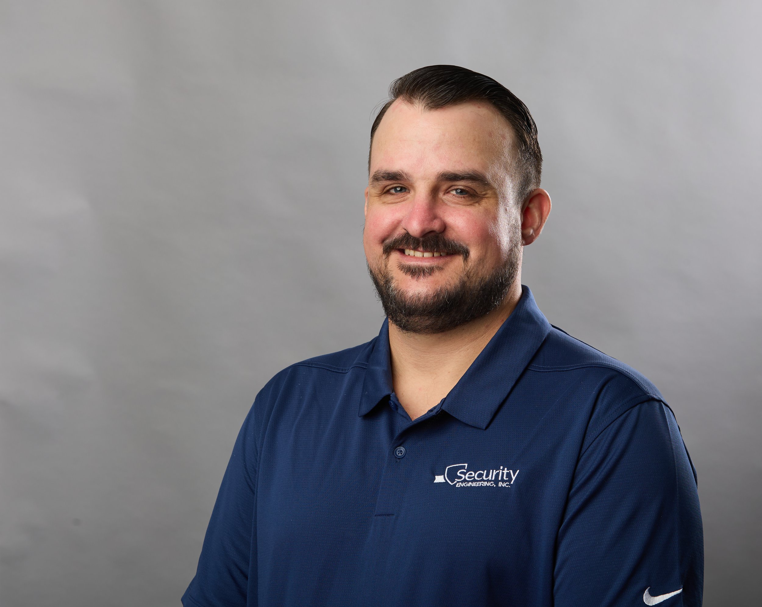Man with short dark hair and beard smiling, wearing a navy blue polo shirt with a 'Security Engineering, Inc.' logo, against a plain gray background.
