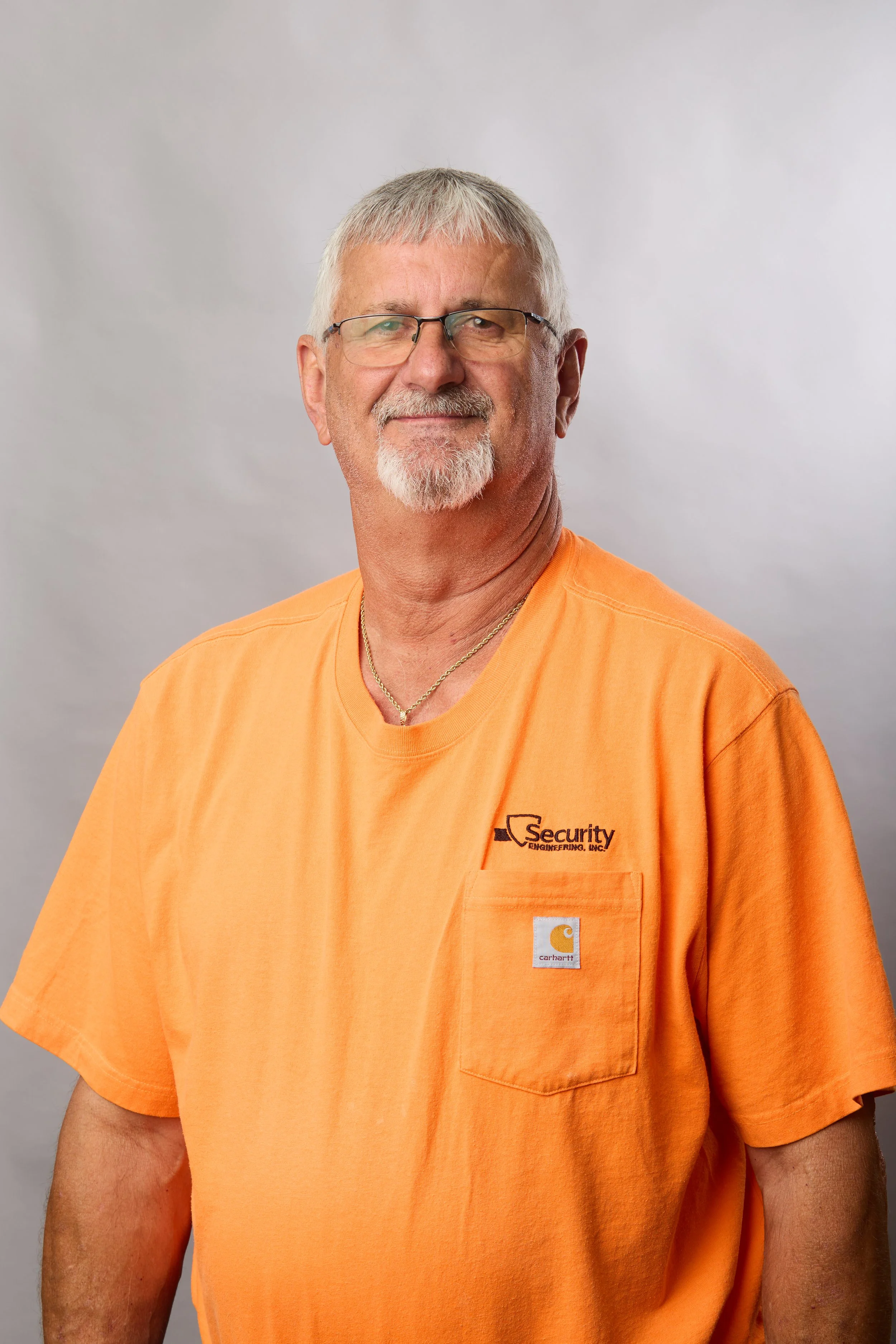 A man with gray hair, glasses, and a goatee wearing an orange T-shirt with company logos and a gold chain, looking at the camera with a neutral expression against a plain backdrop.