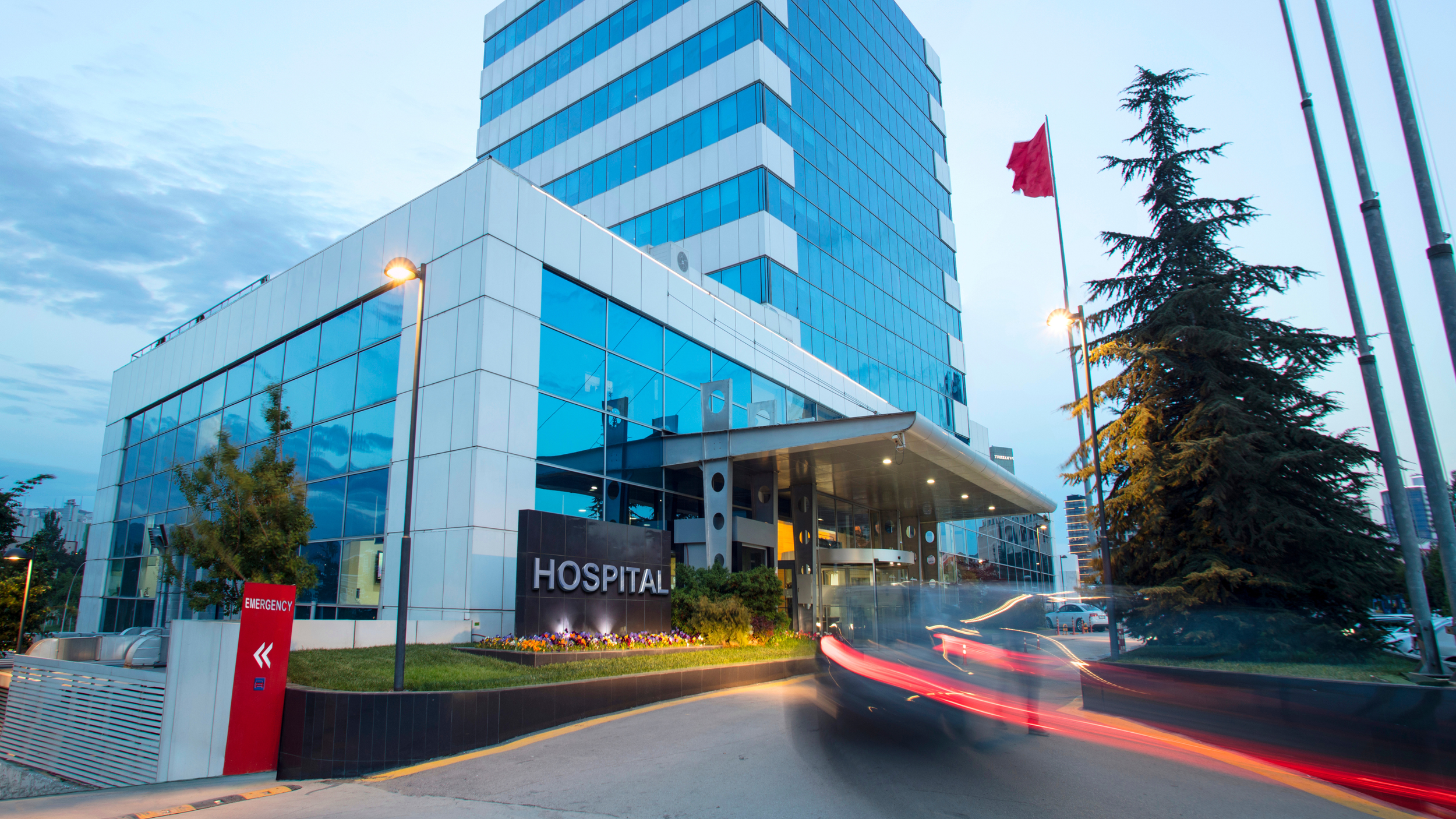 Modern hospital building with glass exterior, a sign reading 'HOSPITAL', an emergency entrance, and a large tree in front, during dusk with light trail from moving vehicles.