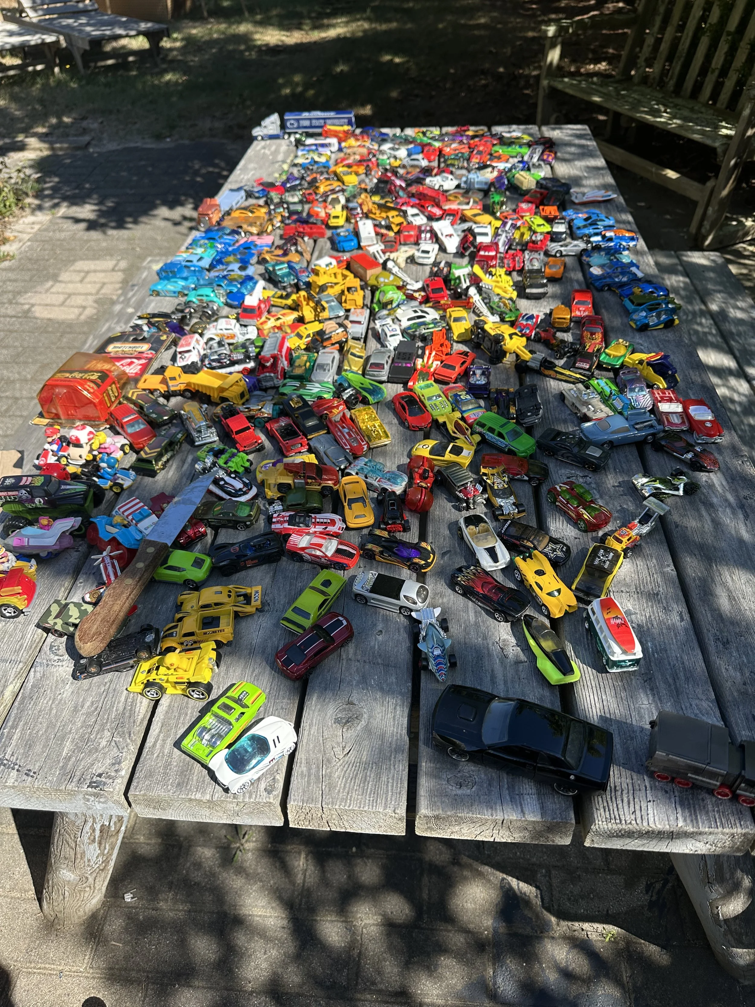 A large collection of toy cars arranged on a weathered wooden table outdoors, with some toys overlapping and a wooden bench in the background.