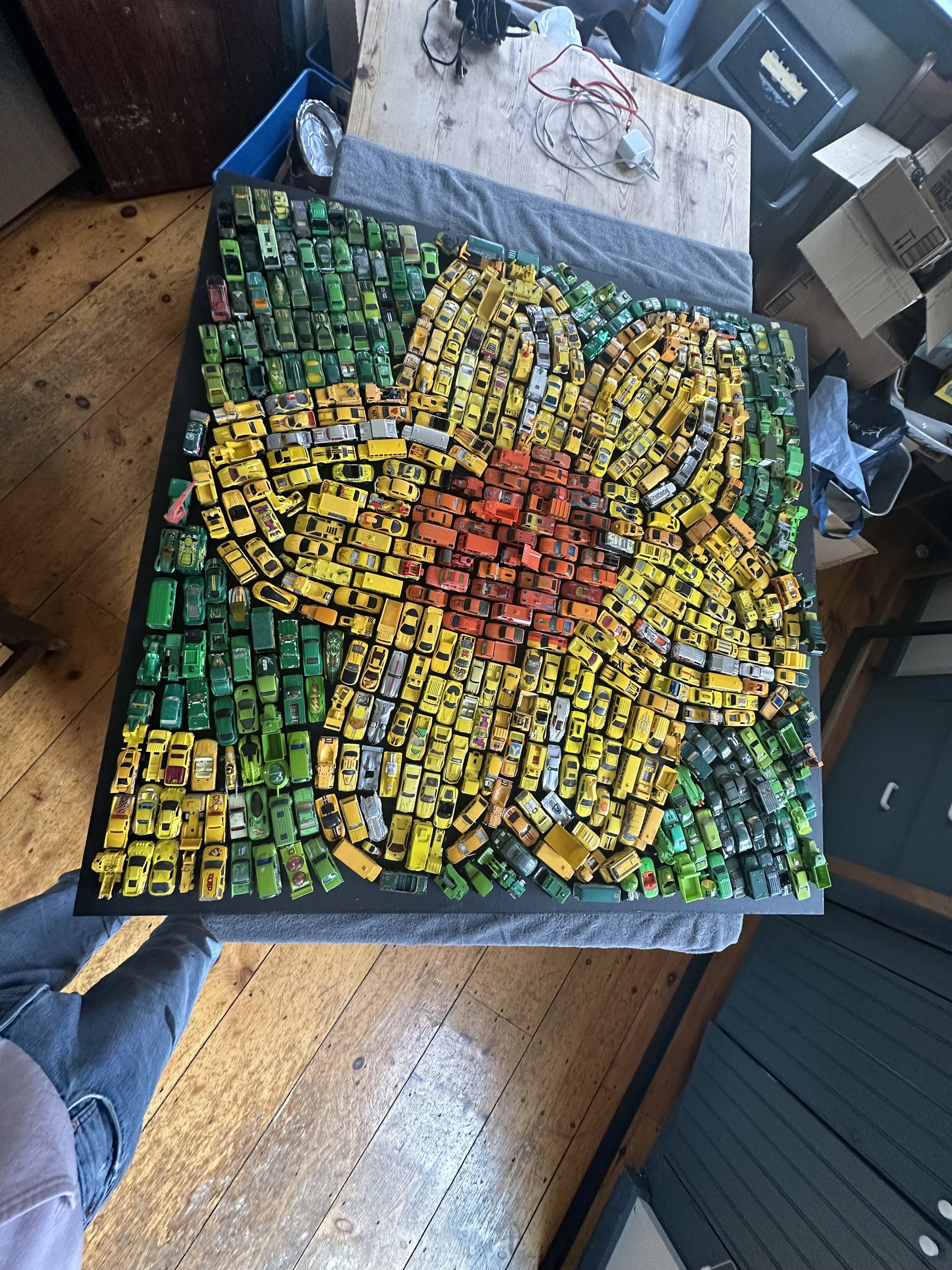 A large collection of small toy cars organized to form a mosaic of a yellow and orange sunflower on a black table.