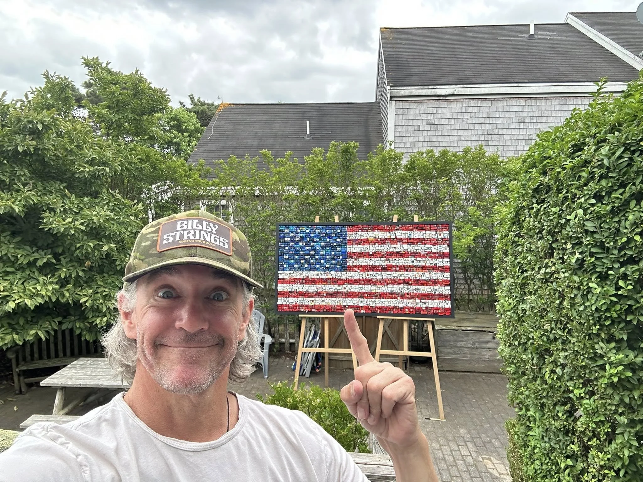 A smiling man with gray hair wearing a camouflage baseball cap that reads 'Billy Strings'. He is pointing to a mosaic American flag made of small tiles on an easel in a backyard, with trees, bushes, and houses in the background.