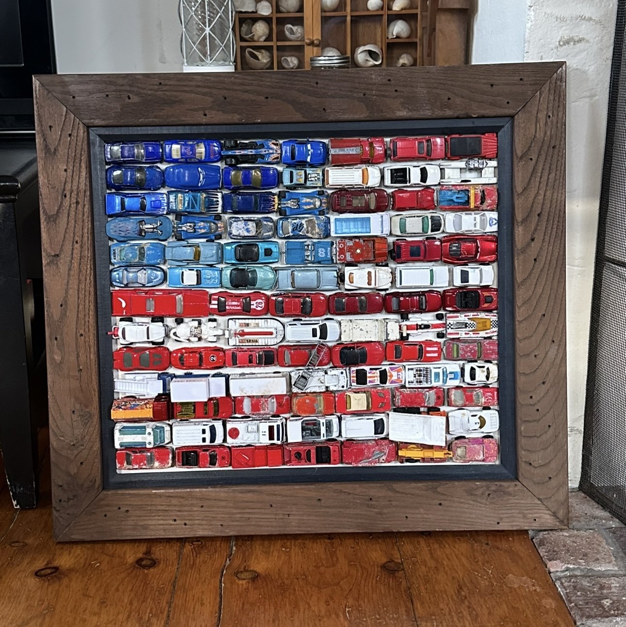 A shadow box frame filled with a collection of miniature toy cars arranged in rows, with a mix of blue, red, white, and other colored cars.
