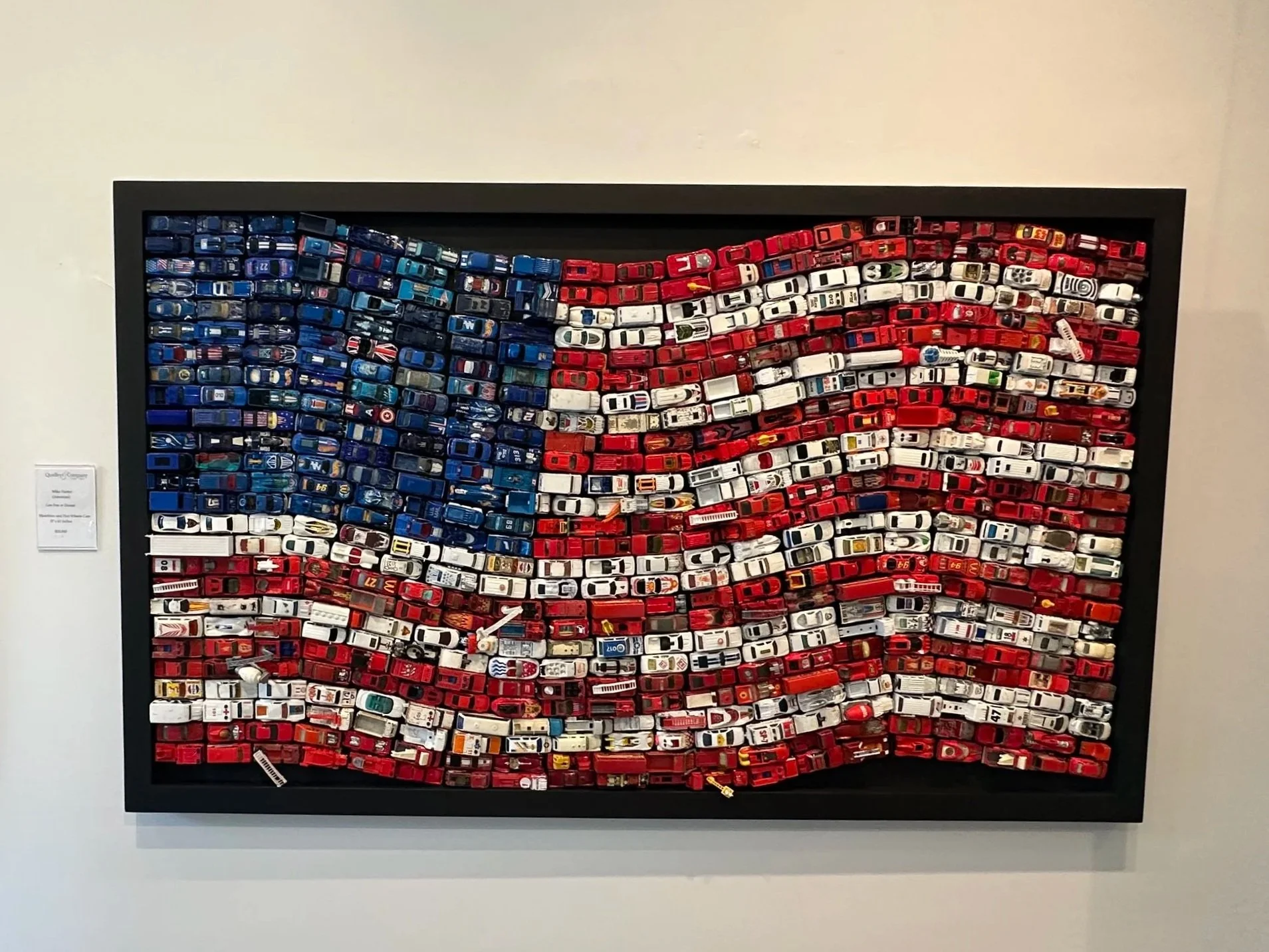 An art piece made of numerous small toy cars arranged to depict the American flag, with blue stars on the left and red and white stripes across the rest of the frame.