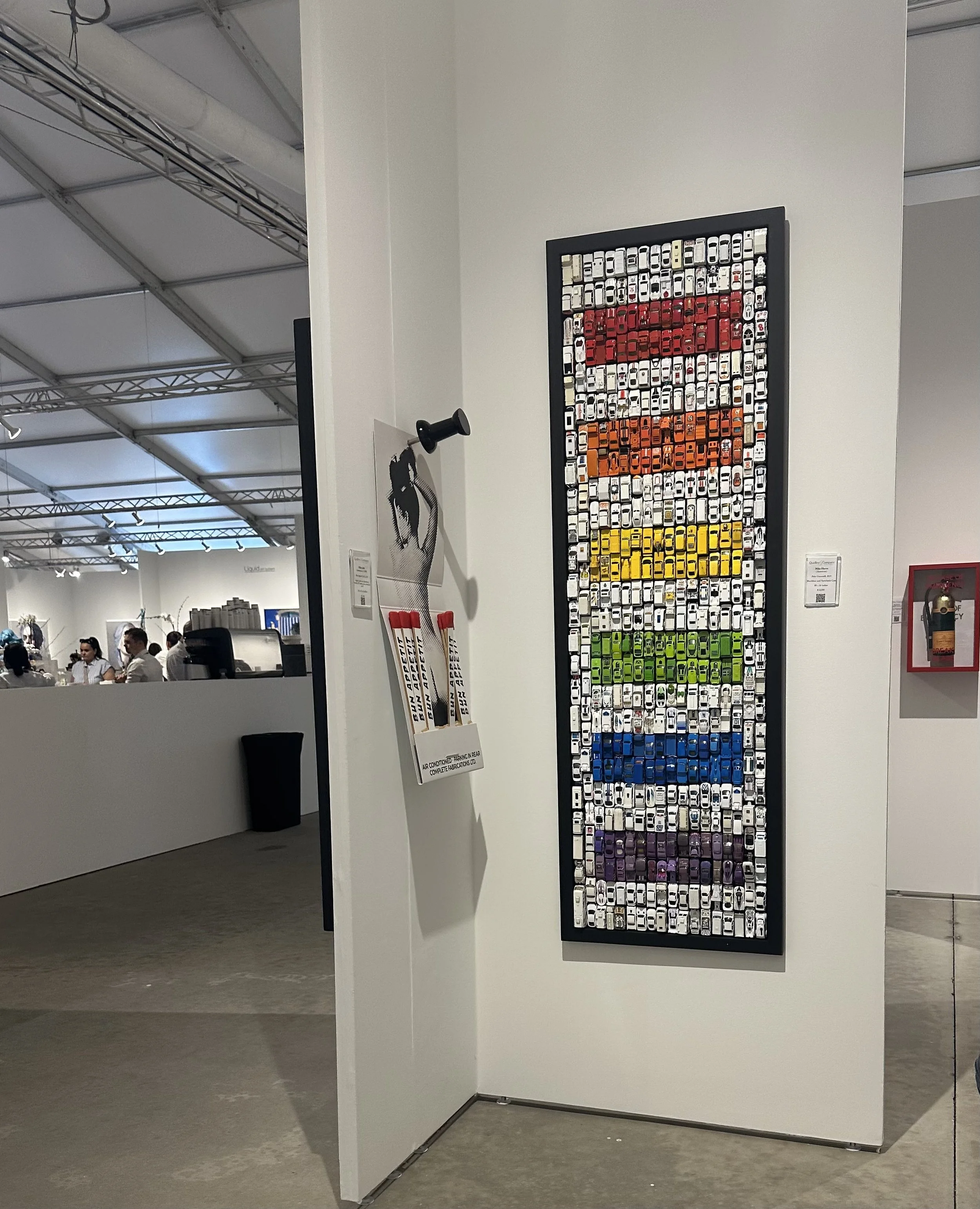 A framed artwork made of tiny car images organized by color in horizontal stripes of red, orange, yellow, green, blue, and purple, displayed on a white gallery wall.