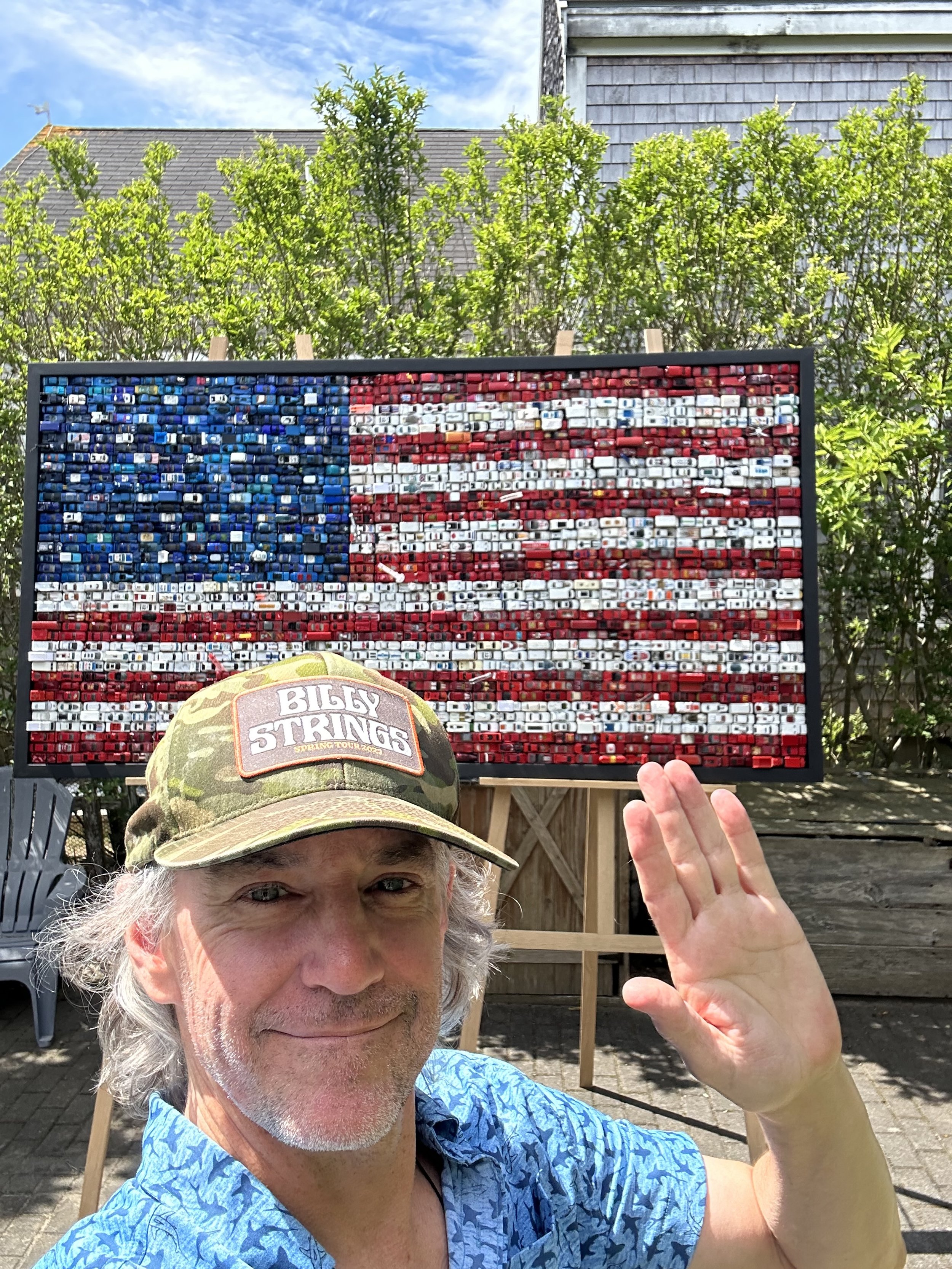 A man with gray hair and a beard, wearing a camouflage cap and a blue patterned shirt, smiling and waving in front of a mosaic American flag made of small bottle caps on an outdoor easel.
