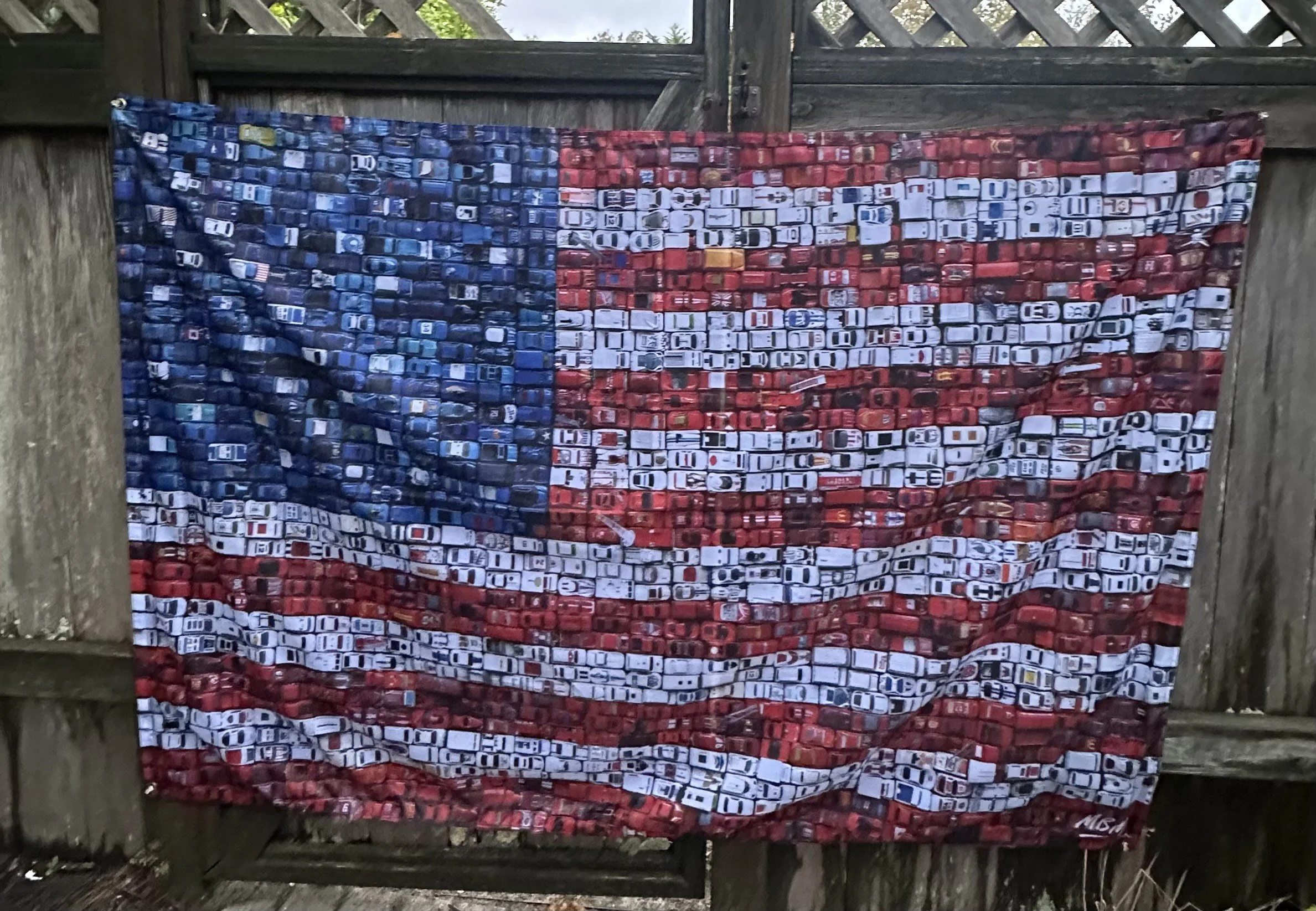 A quilt designed to resemble the American flag with stars and stripes made from small fabric squares, hanging on a wooden fence.