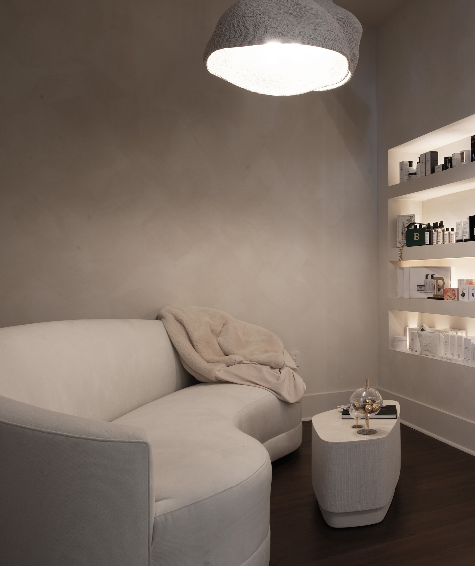 A cozy corner with a white curved sofa, a beige fuzzy blanket draped over it, a small matching white table with a glass bowl, and built-in shelves with skincare products and decor, illuminated by a large hanging lamp.