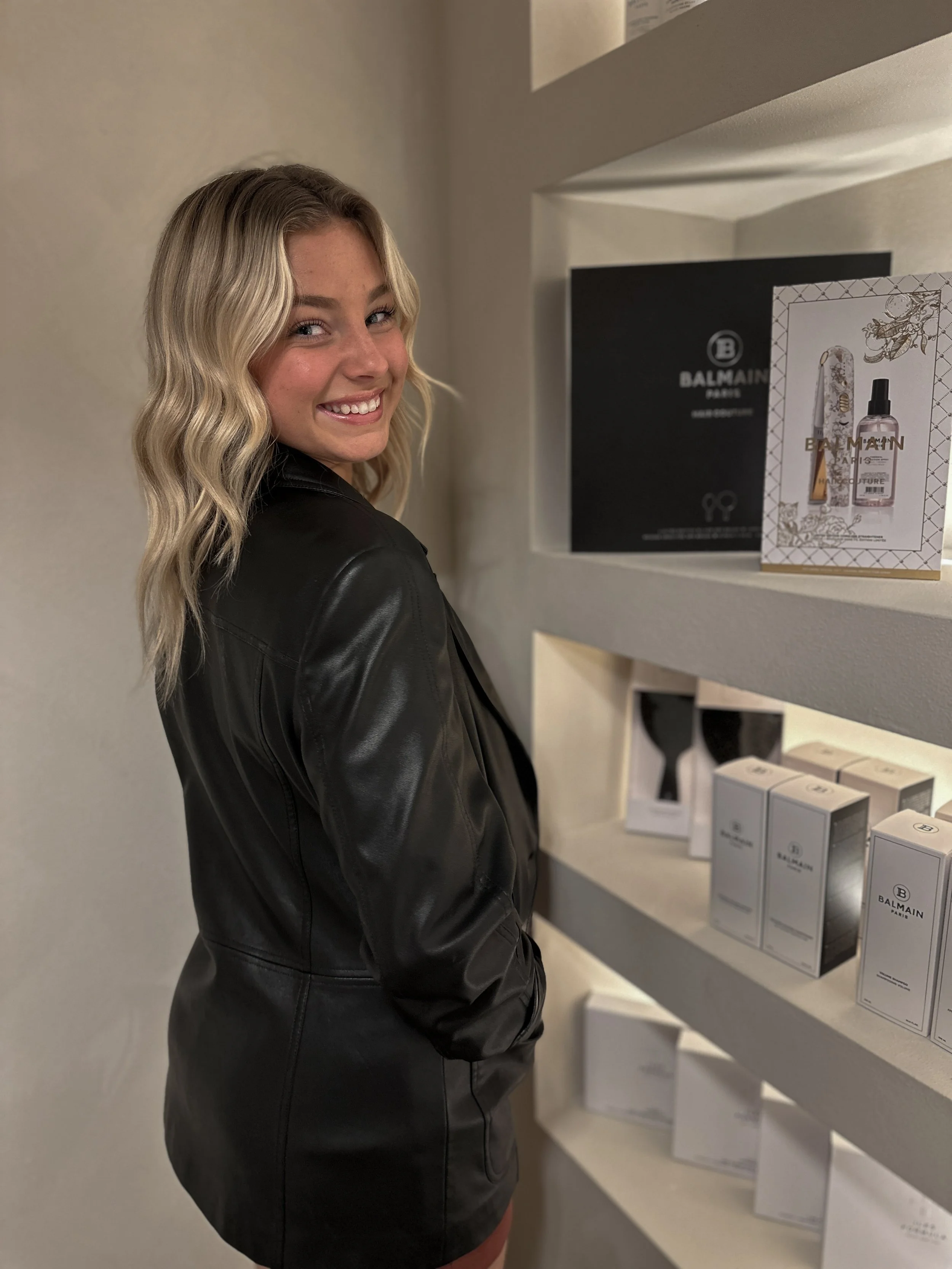 A smiling young woman with wavy blonde hair, wearing a black leather jacket, standing in front of shelves displaying Balmain Paris products.