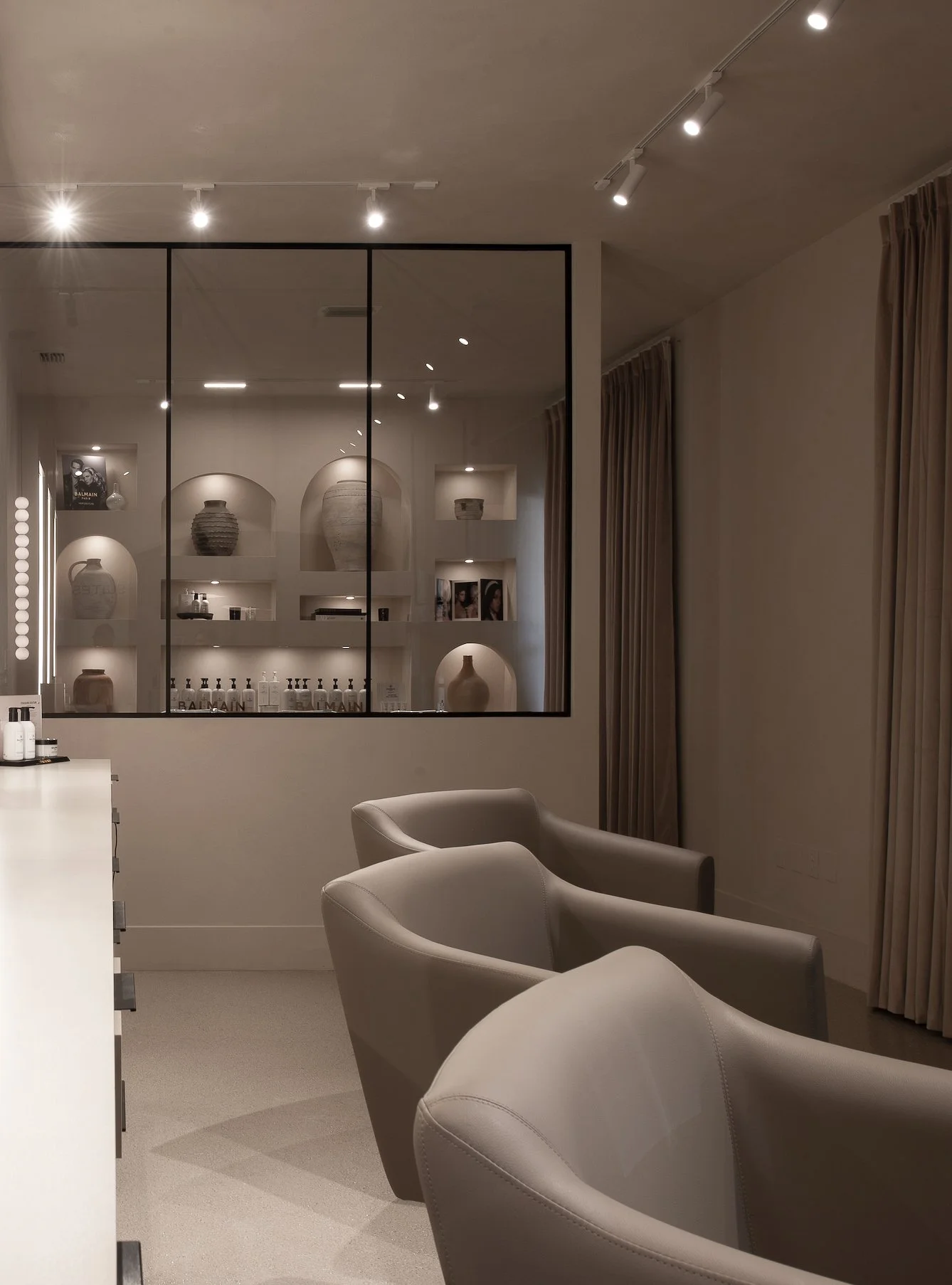 Modern interior space with three beige armchairs in the foreground, glass partition with shelves and decorative vases, and curtains on the right wall, illuminated by track lighting.