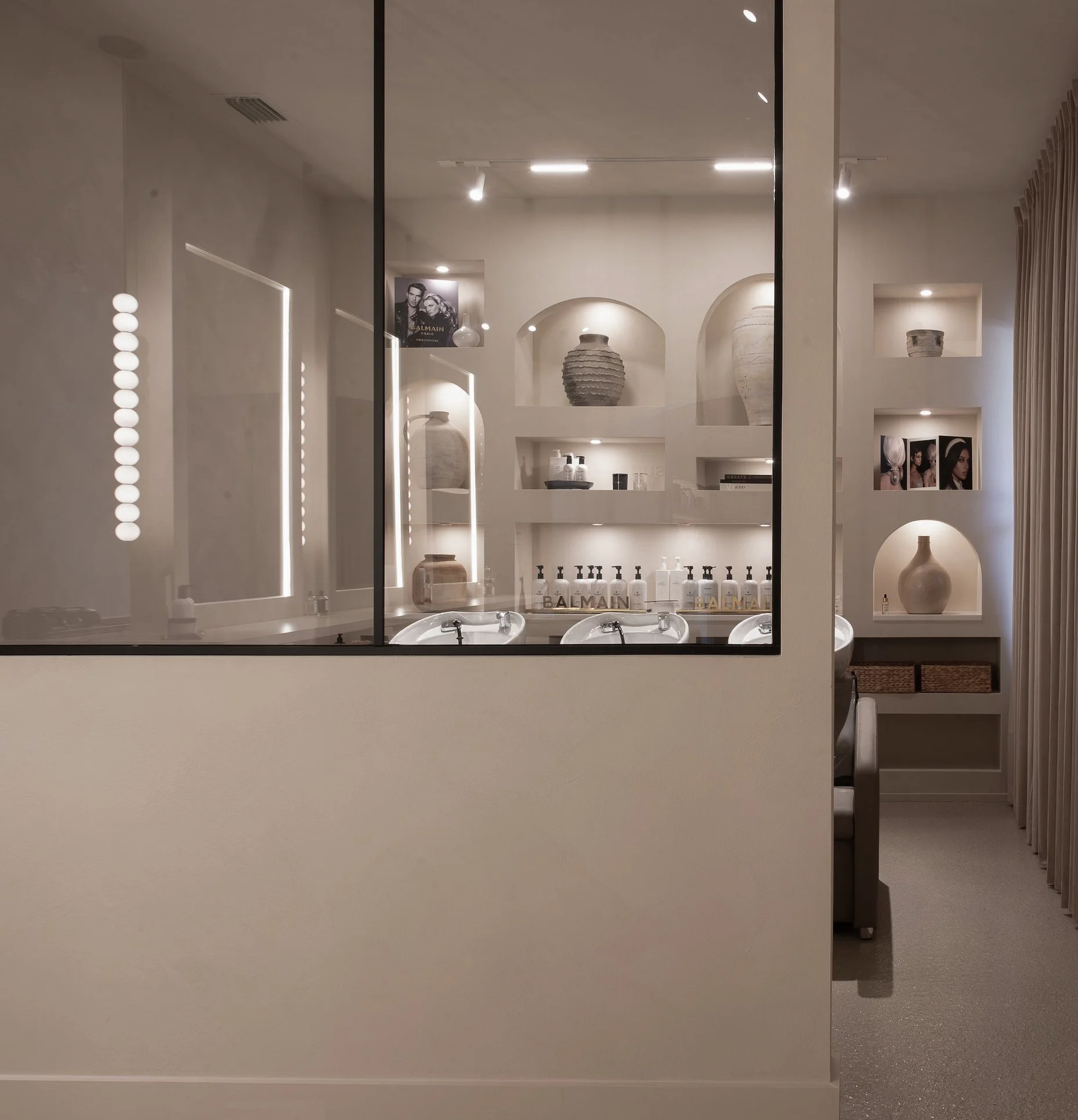 A glimpse through a glass partition into a modern, minimalist bathroom with white walls, decorative shelves with vases, personal care products, framed photos, and a vanity with sinks.