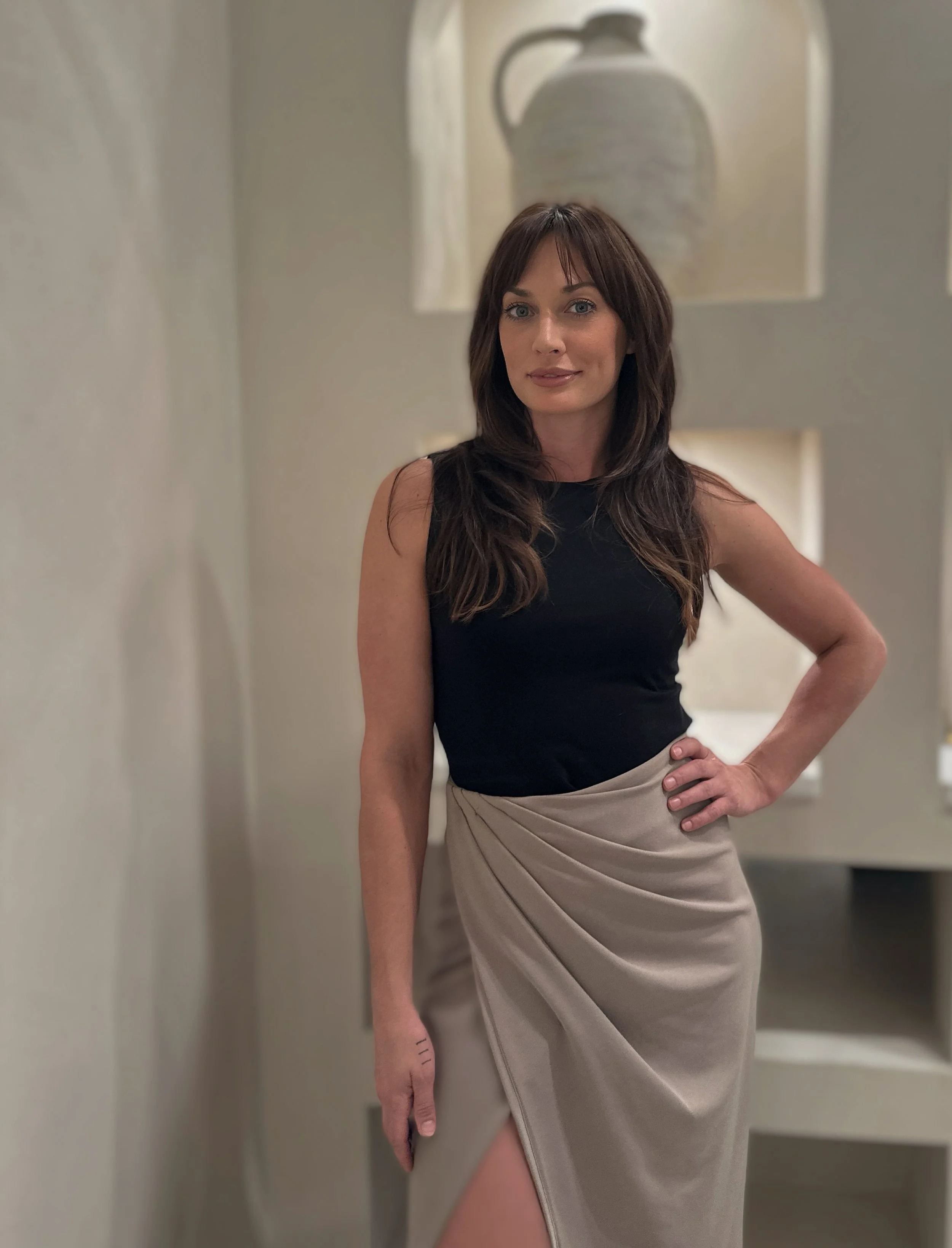 A woman with long brown hair wearing a black sleeveless top and a beige skirt with a slit, standing indoors with one hand on her hip.