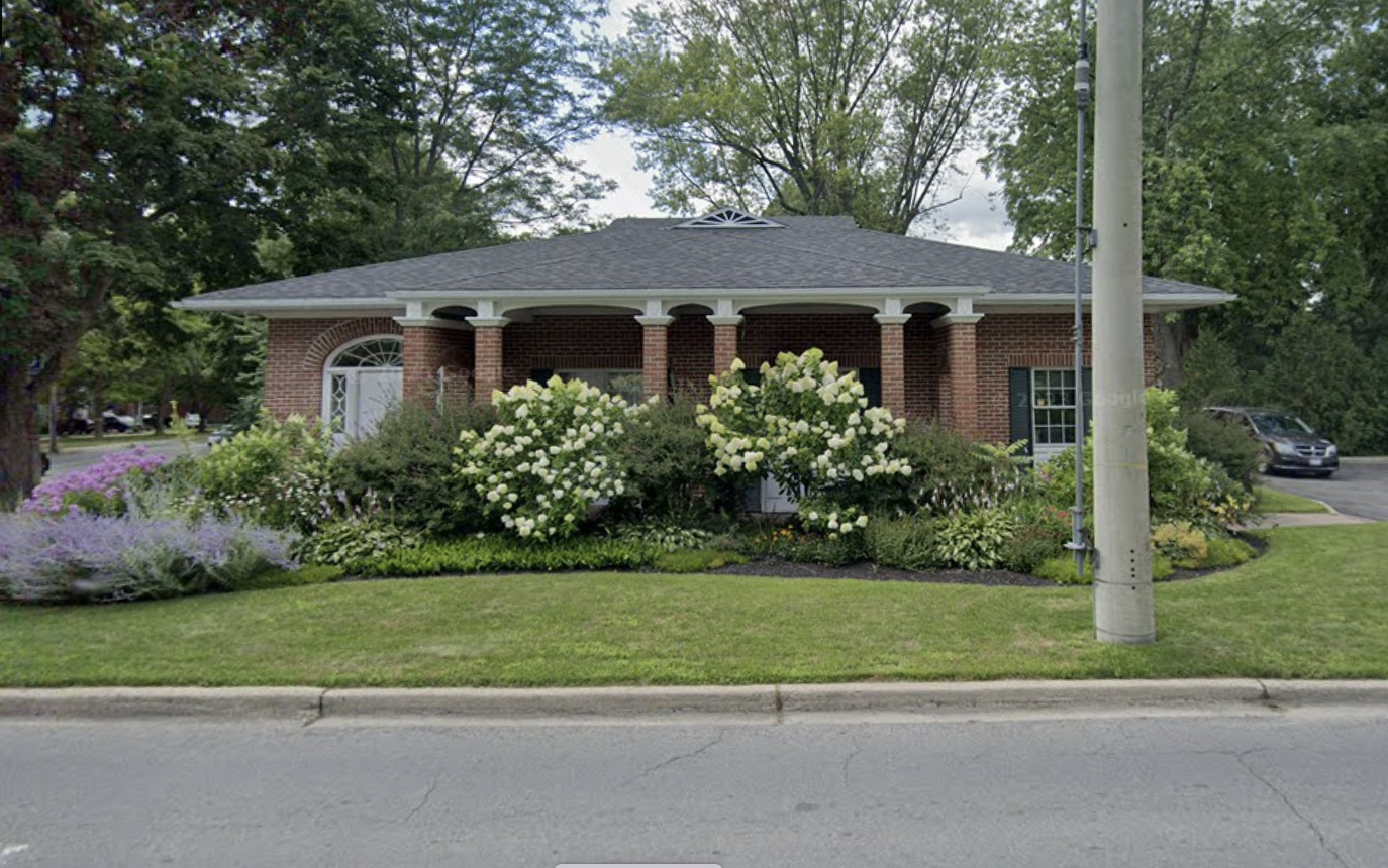 A brick building with a grey shingled roof, surrounded by landscaped bushes and flowering plants. There is a sidewalk in front and a parking lot with cars to the right. A utility pole is also visible.